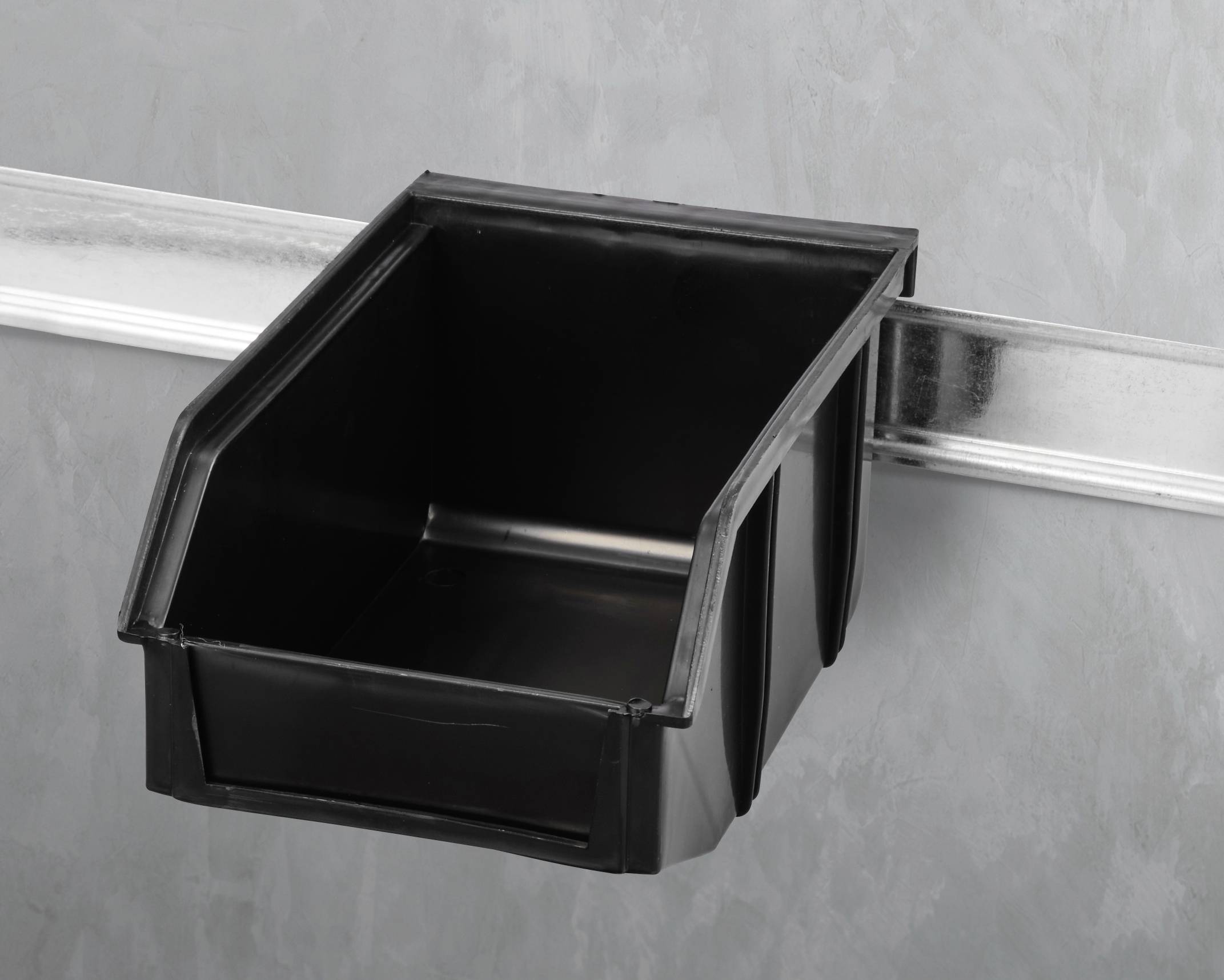 Ein schwarzer Kunststoffbehälter hängt an einer Metallschiene an der Wand. Der Behälter ist leer und hat eine offene Seite.