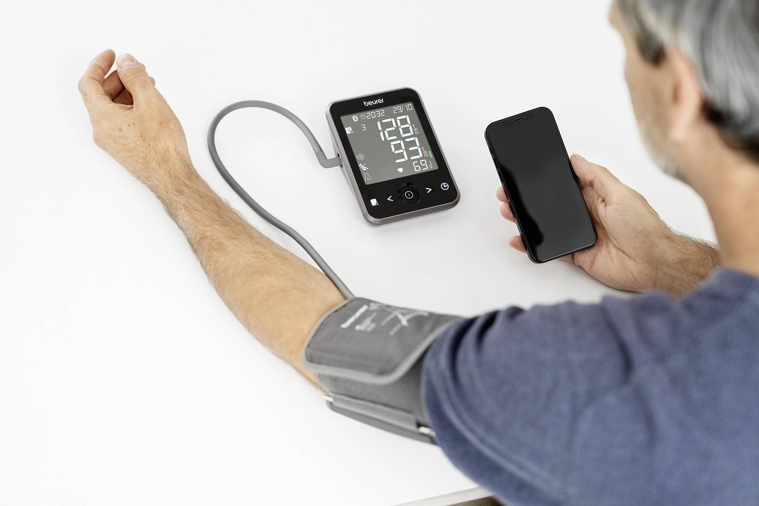 Ein Mann misst seinen Blutdruck mit einem digitalen Blutdruckmessgerät, während er ein Smartphone in der anderen Hand hält.