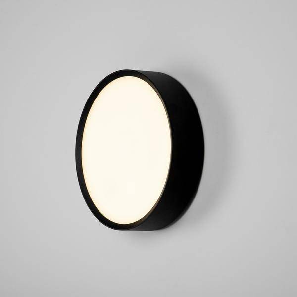 Runde Wandleuchte mit schwarzem Rahmen, leuchtet gleichmäßig. Simples, modernes Design auf grauem Hintergrund.
