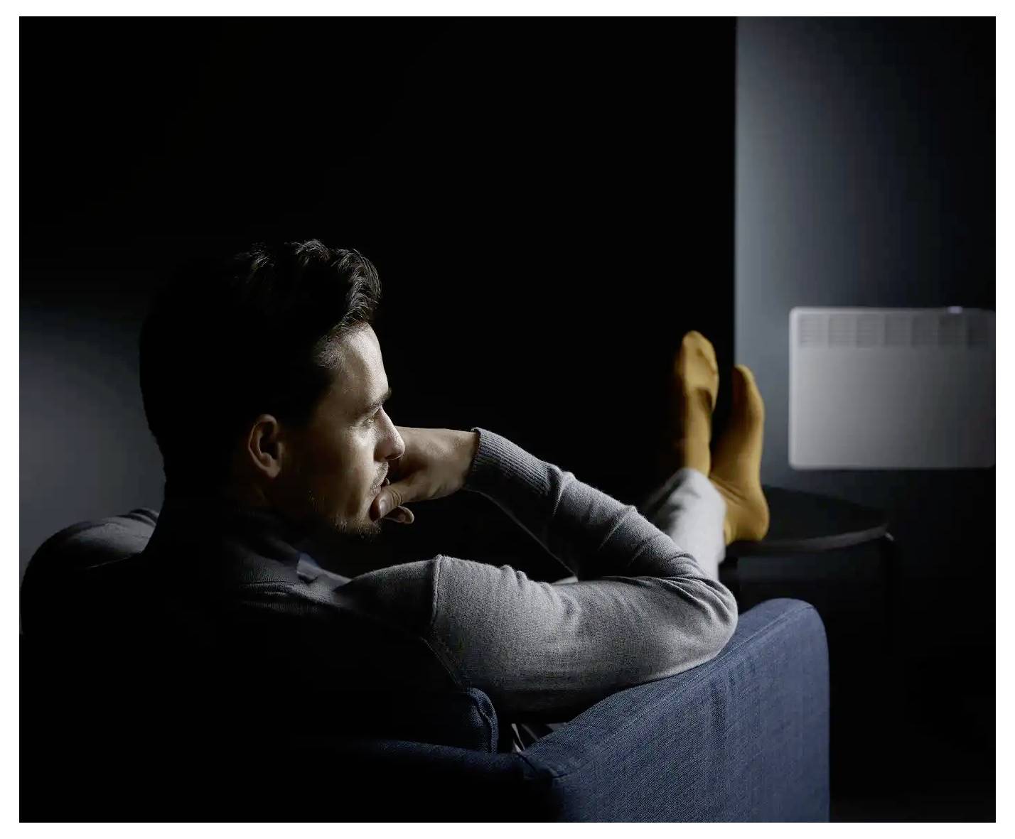 Eine Person sitzt auf einem Sofa in einem schwach beleuchteten Raum, wirkt nachdenklich, trägt orange Socken und wird von einem kleinen weißen Heizgerät an der Wand beleuchtet.