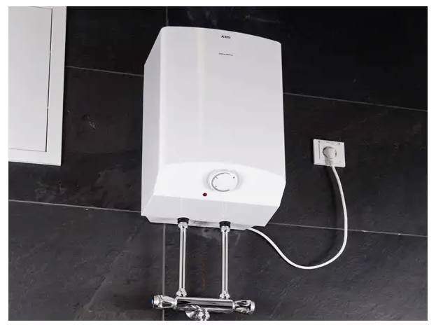 Ein weißer, an der Wand montierter elektrischer Warmwasserspeicher mit einer Skala und roter Anzeigelampe ist in eine Steckdose an einer dunklen Fliesenwand eingesteckt.