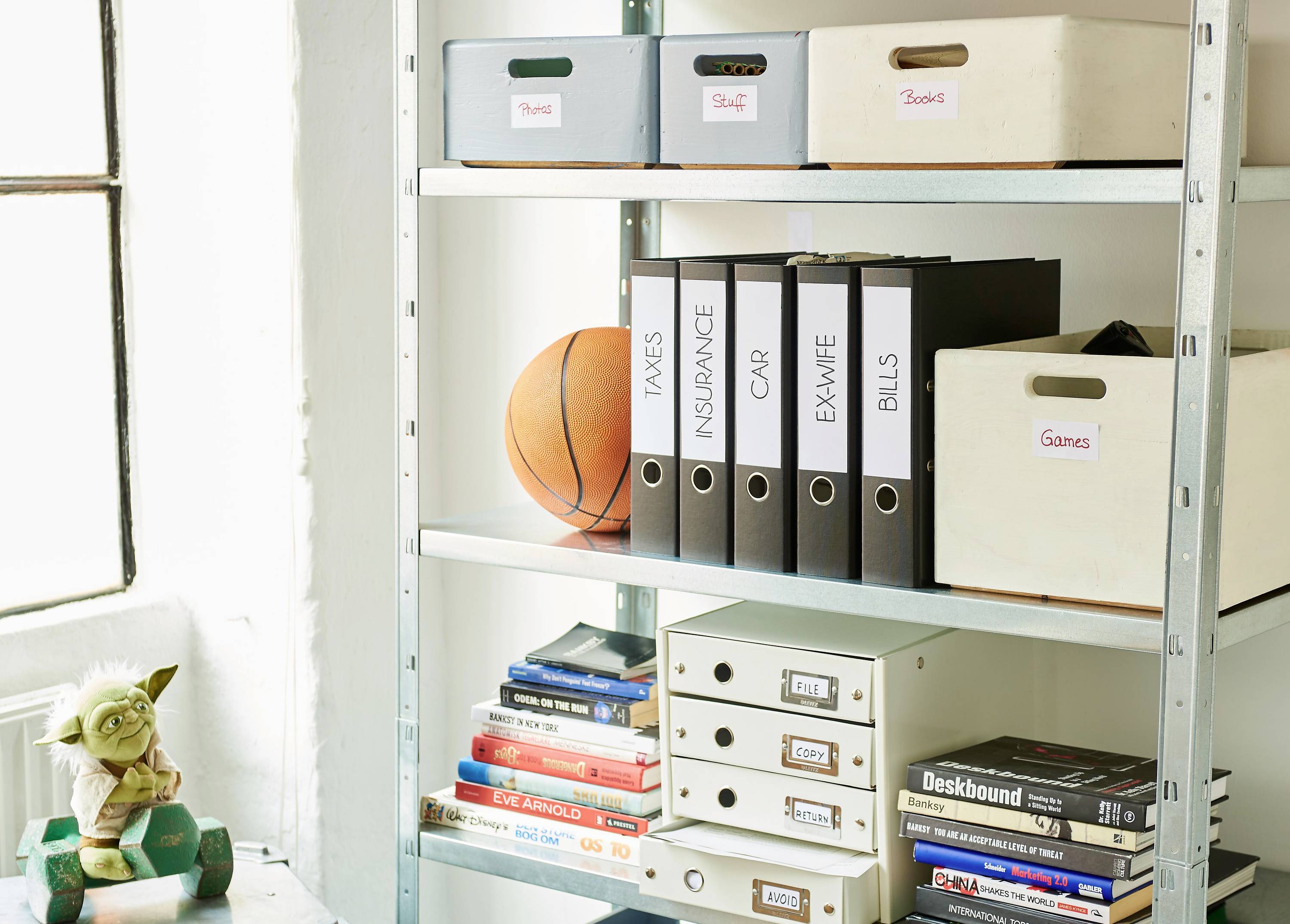 Ein Regal mit Ordnern, beschriftet mit 'Taxes', 'Insurance', 'Car', 'Exwife Bills', sowie Boxen, Büchern und einem Basketball.