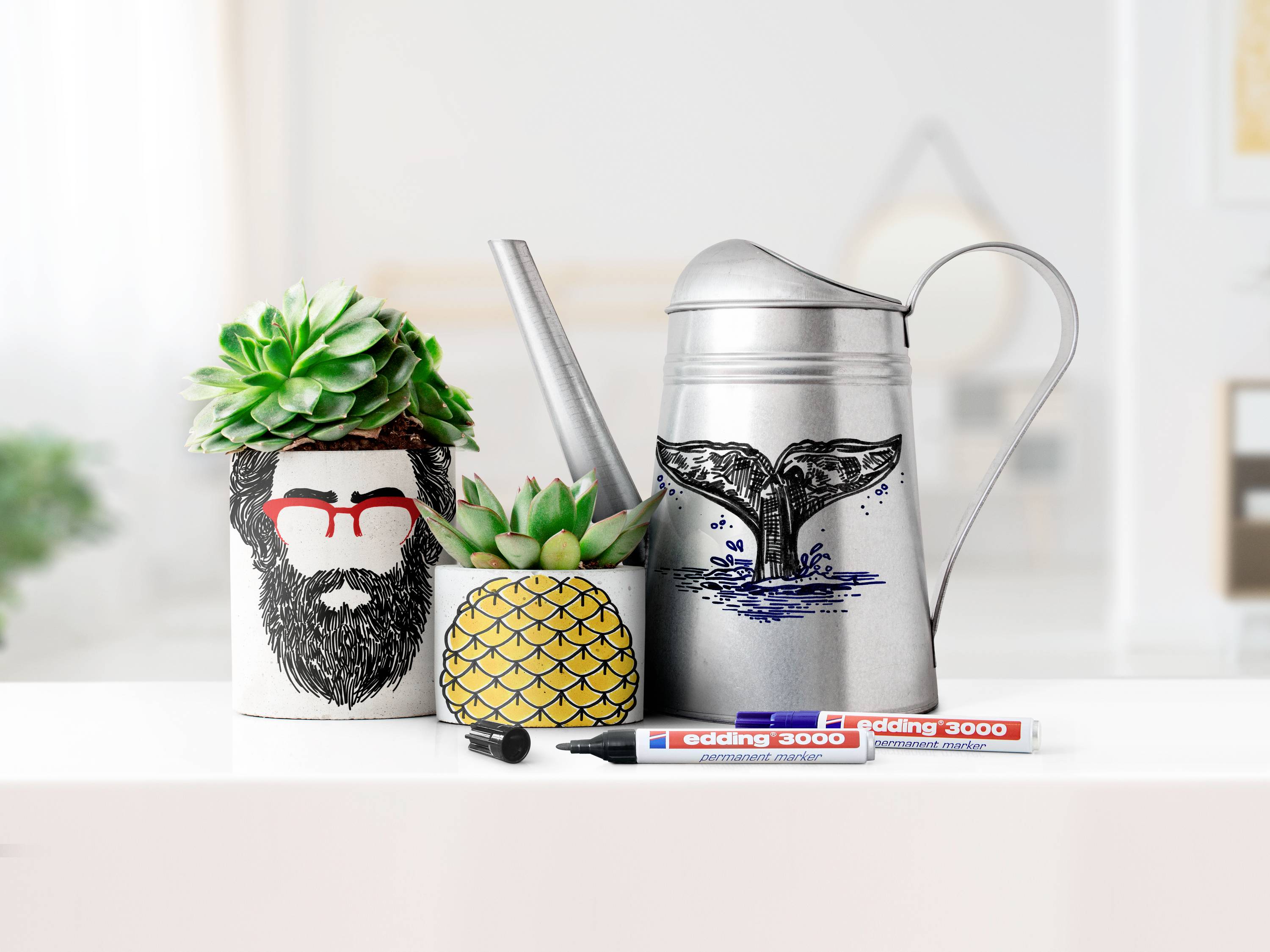 Eine Gießkanne mit Walflossen-Design steht zwischen einem Ananas-Topf und einem dekorierten Pflanzgefäß. Vorne liegen zwei Marker.