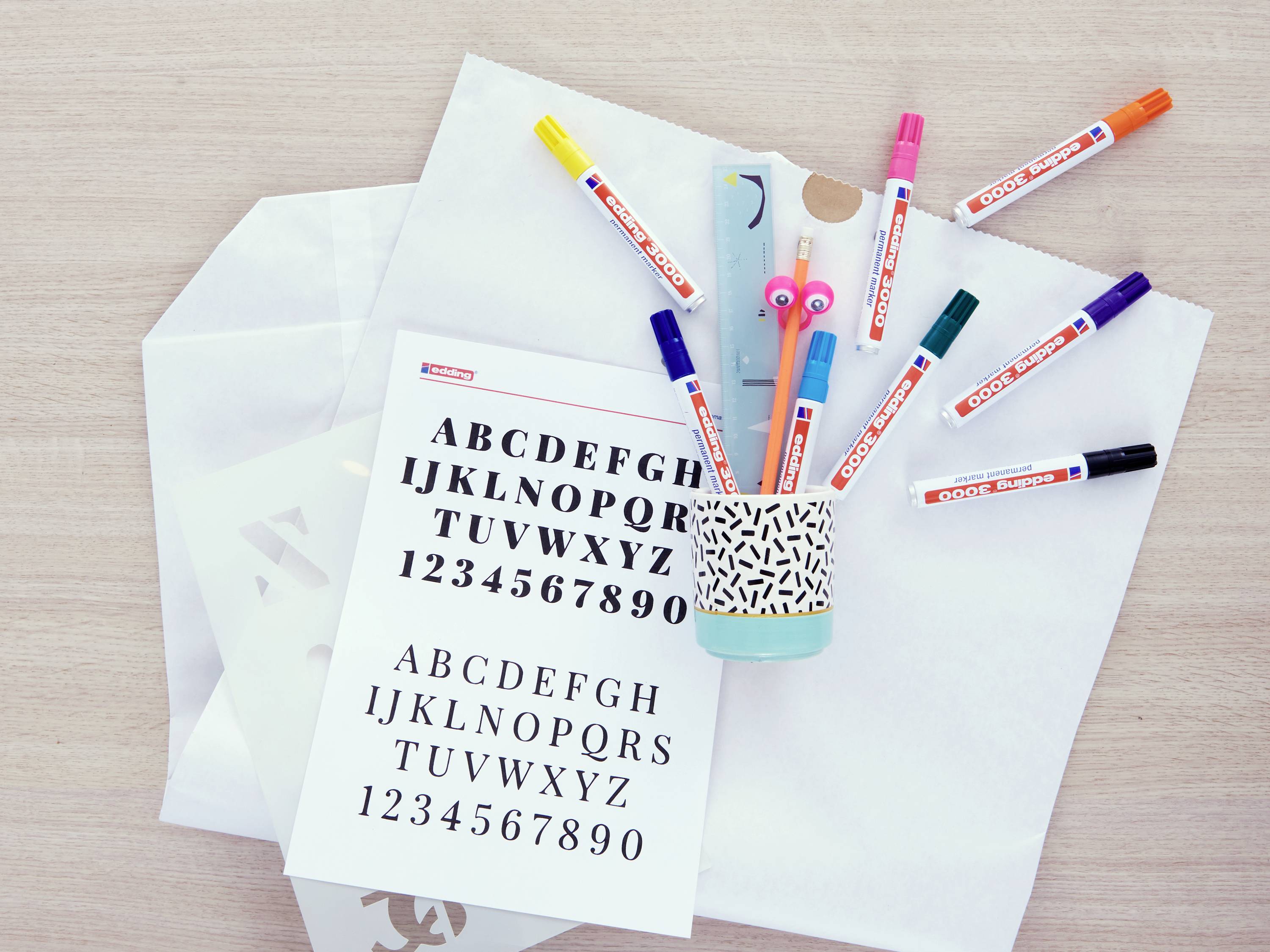 Stifte in einem Becher, eine Vorlage mit Alphabet und Zahlen auf einem Tisch. Schreib- und Bastelmaterialien liegen bereit.