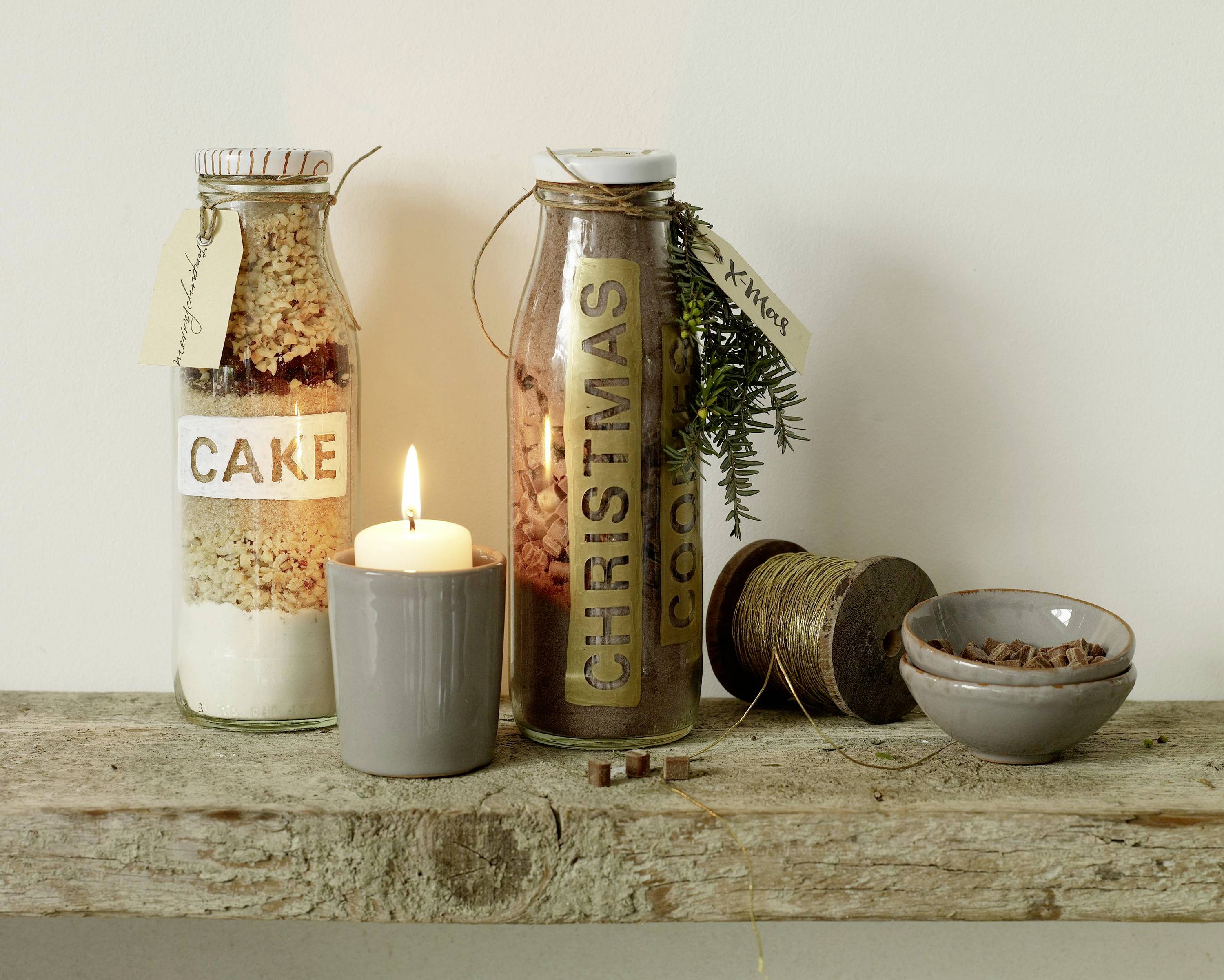 Zwei Gläser mit Zutaten für Kuchen und Kekse, beschriftet mit 'Cake' und 'Christmas Cookies', stehen auf einem Holzregal neben einer brennenden Kerze.