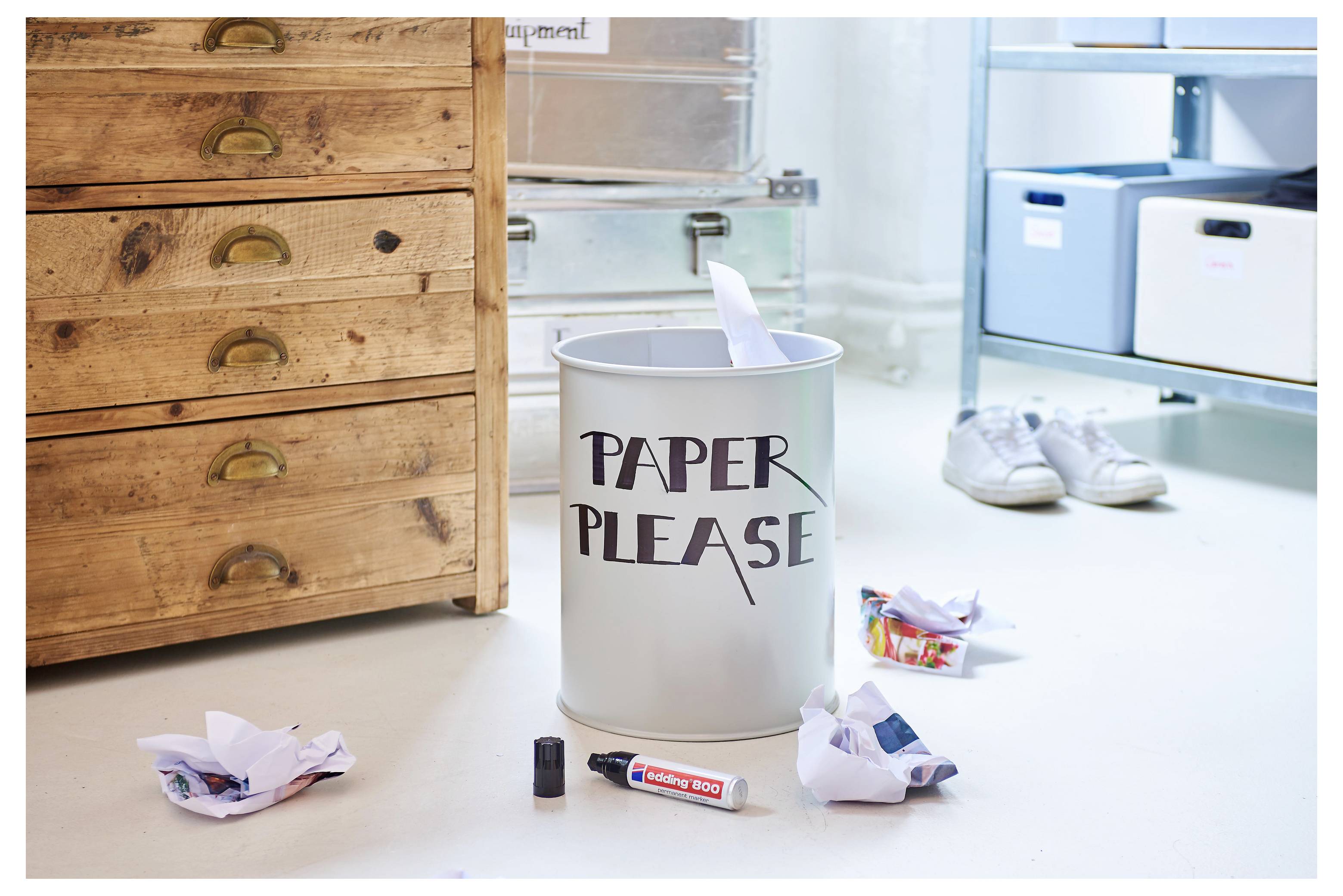 Papierrecyclingbehälter mit der Aufschrift "PAPER PLEASE" in einem Raum, umgeben von zerknittertem Papier, mit Schuhen und Lagerschubladen im Hintergrund.