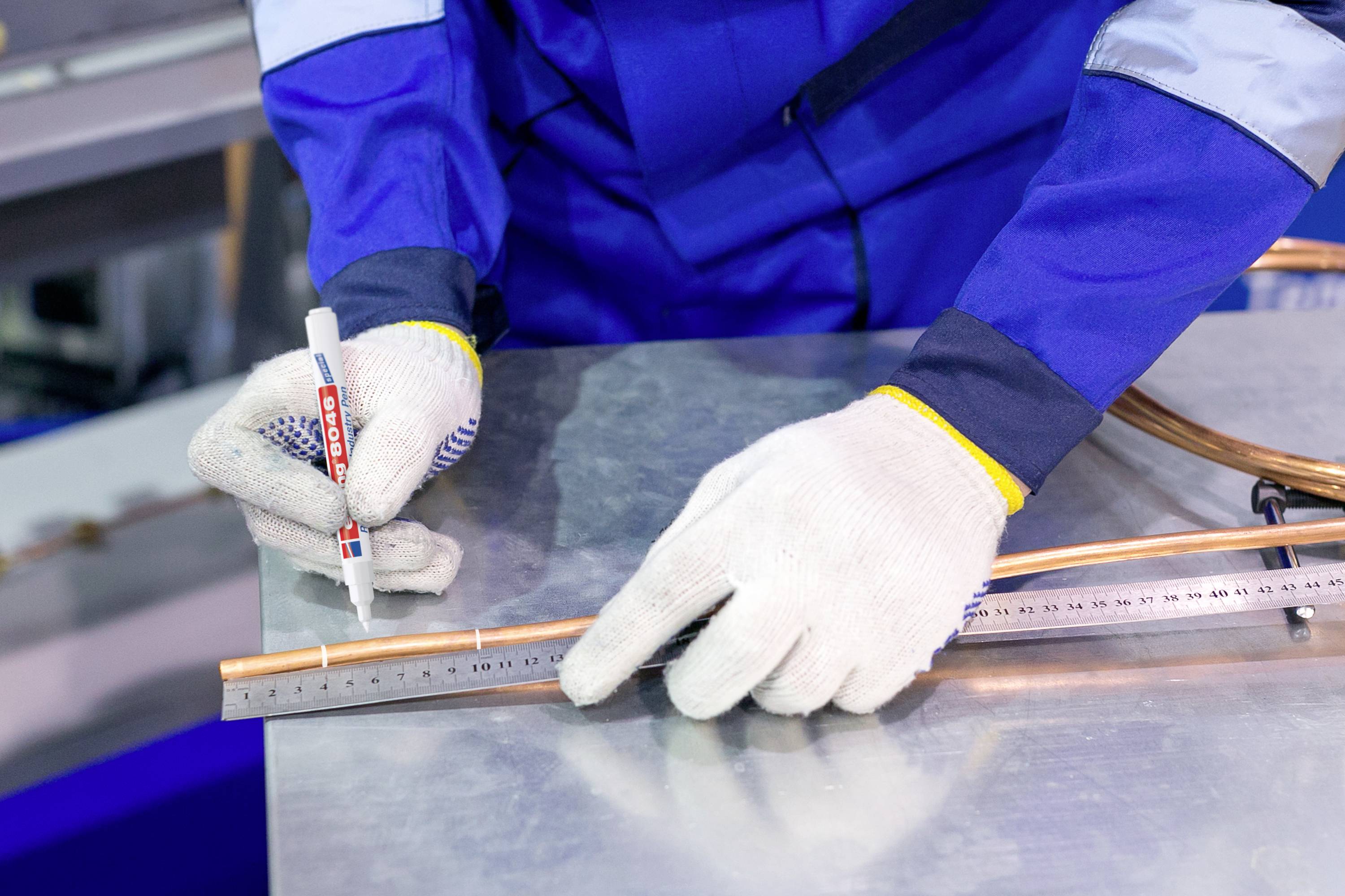 Eine Person in blauer Arbeitskleidung und Handschuhen misst ein Kupferrohr mit einem Lineal und markiert es mit einem Stift.