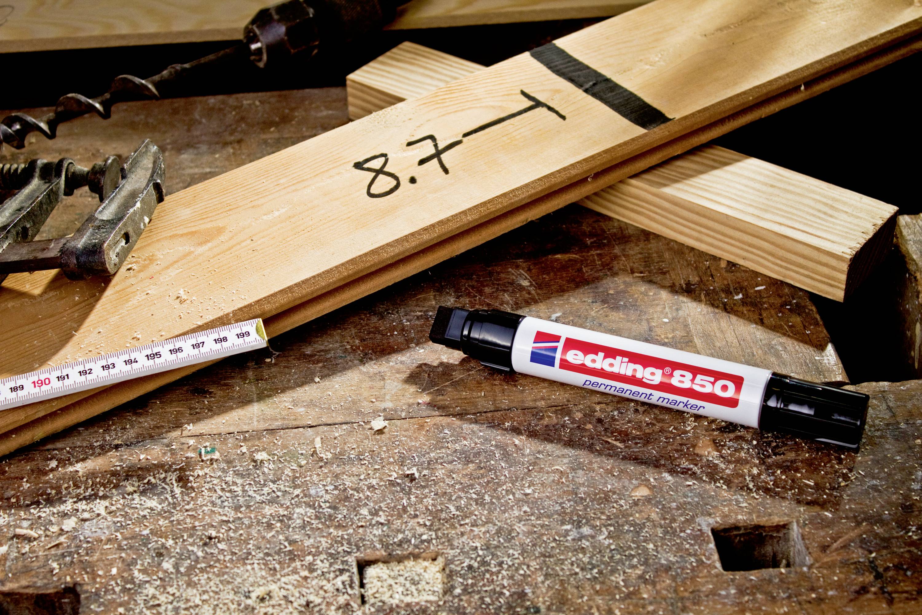 Holzbrett auf Werkbank mit schwarzer Markierung '8,7', daneben ein Maßband und ein schwarzer Permanentmarker, Marke sichtbar.
