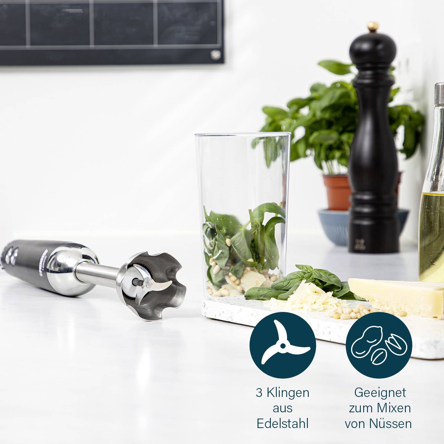 Ein Stabmixer mit Edelstahlklingen liegt auf einem Tisch. Daneben ein Glas mit Kräutern und Nüssen, geeignet zum Mixen von Nüssen.
