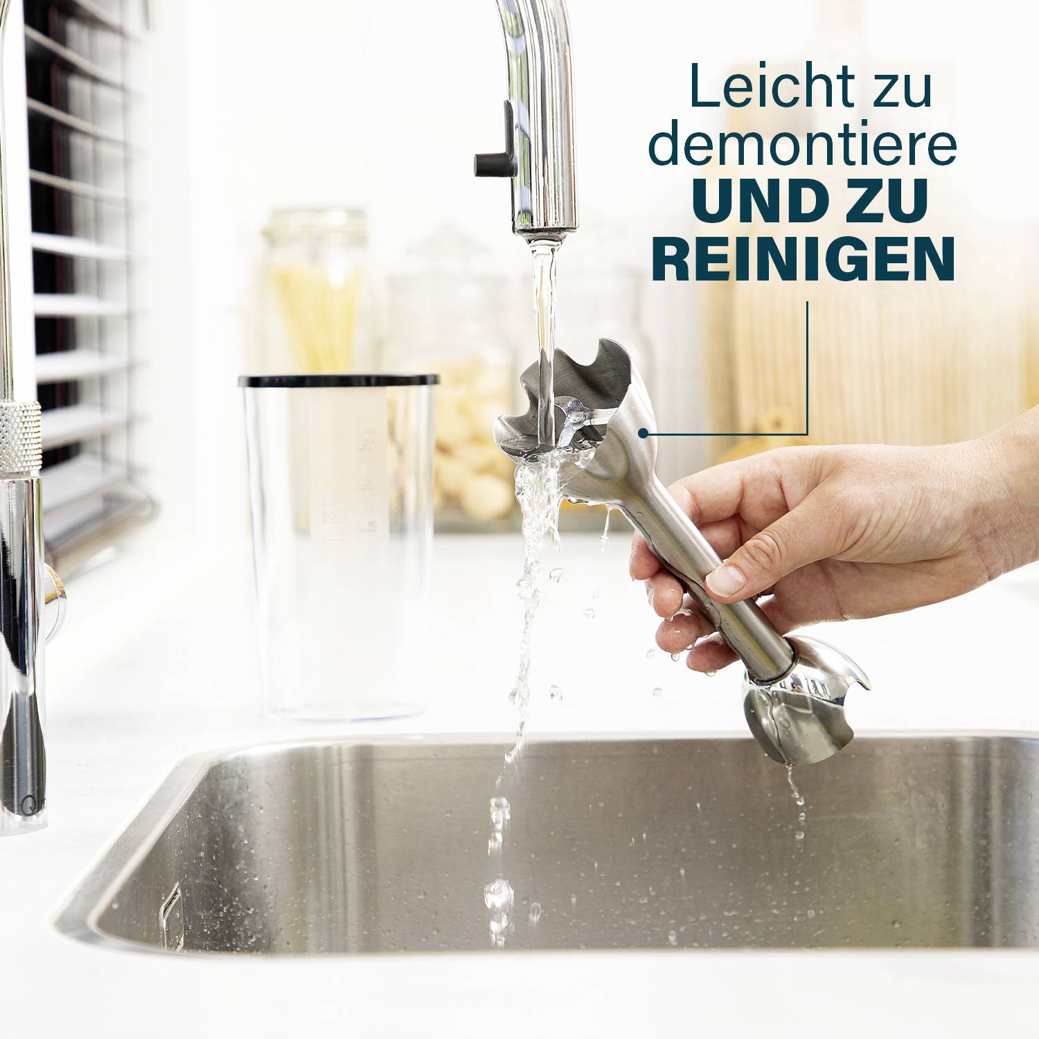 Eine Hand hält einen abnehmbaren Stabmixer-Aufsatz über einem Spülbecken unter fließendem Wasser. Text: 'Leicht zu demontieren und zu reinigen'.