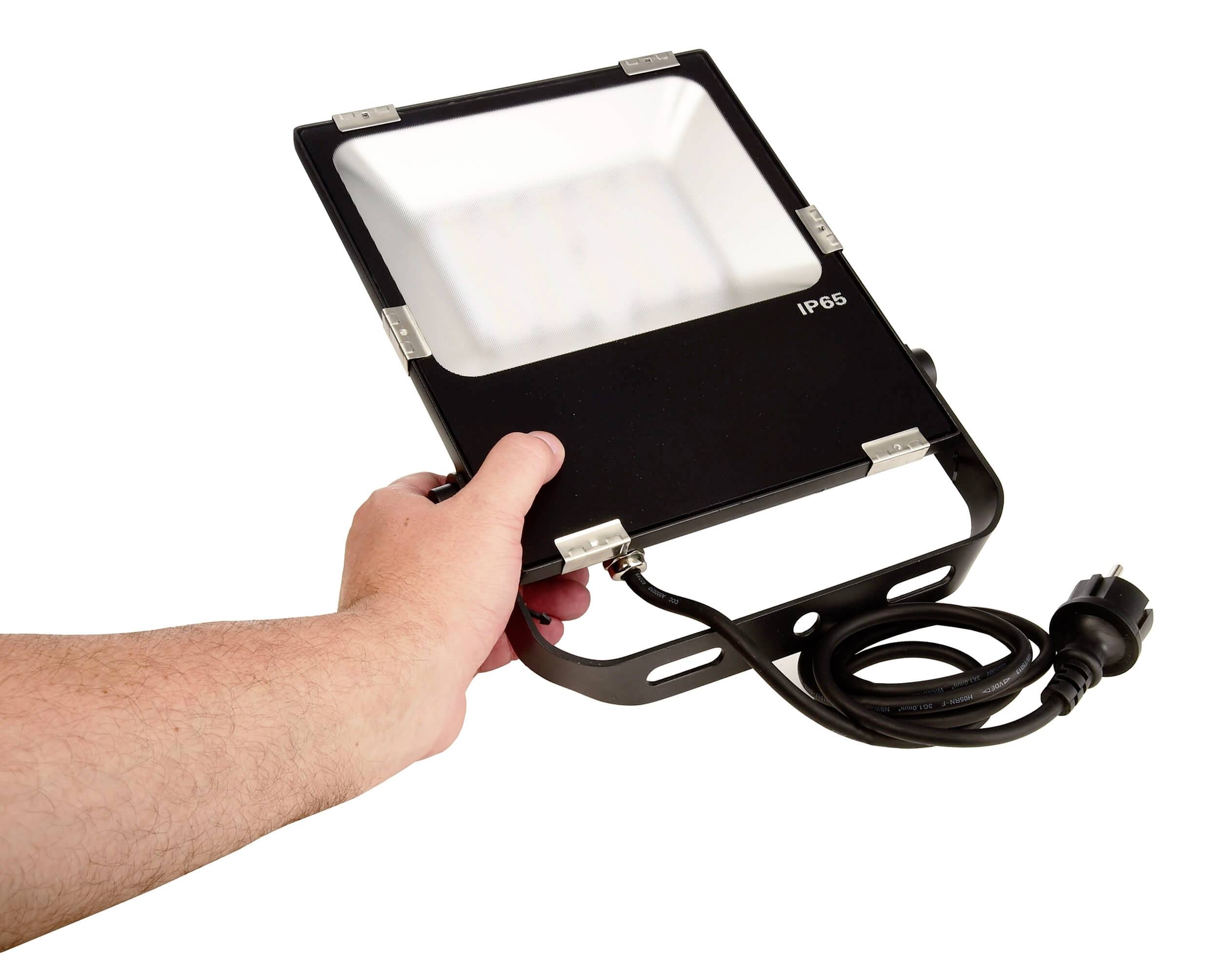 Eine Hand hält einen schwarzen LED-Strahler mit IP65-Schutzklasse. Der Strahler hat ein Stromkabel mit Stecker und eignet sich für den Außeneinsatz.