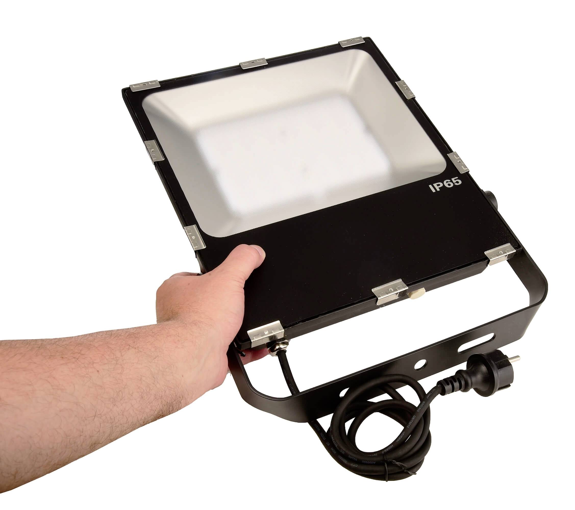 Hand hält einen rechteckigen LED-Scheinwerfer mit Kabel und Stecker. Auf der Vorderseite steht 'IP65'.