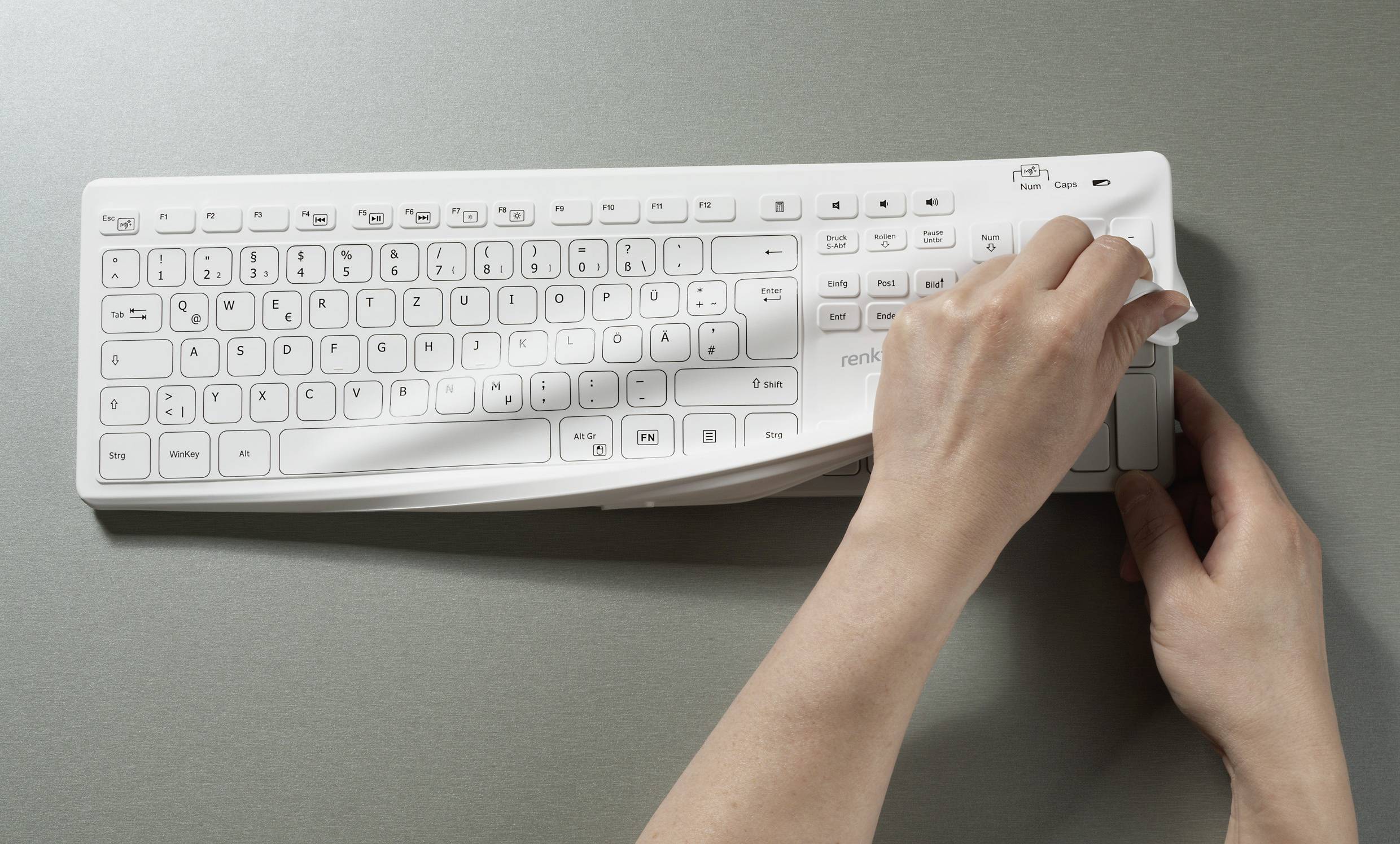 Zwei Hände ziehen eine transparente Schutzfolie von einer weißen Tastatur auf einem grauen Hintergrund ab.