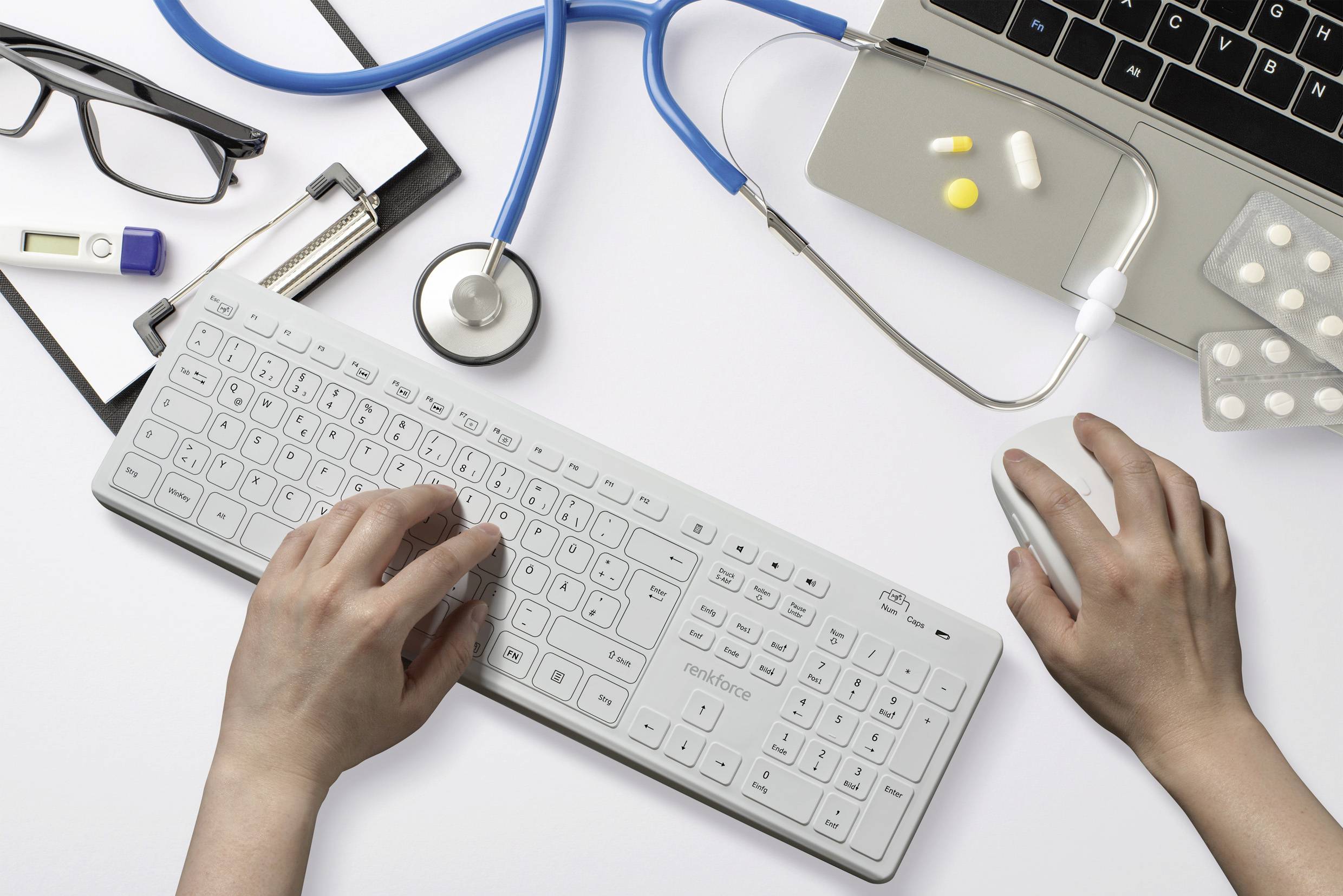 Hände tippen auf Tastatur neben Laptop, Stethoskop und Tabletten, was auf Nutzung von Technologie im medizinischen Bereich hindeutet.