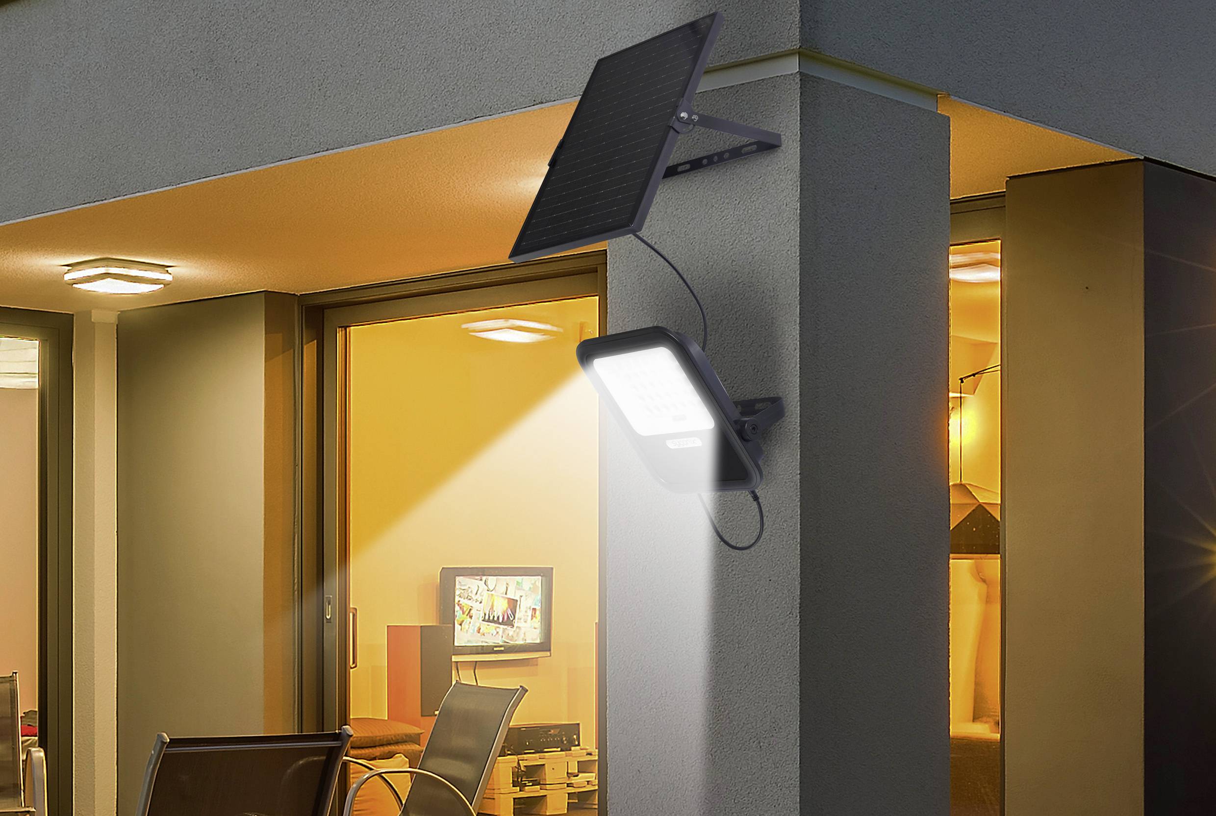 Eine Solarlampe ist an einer Hauswand befestigt und beleuchtet den Bereich darunter. Im Hintergrund ist ein erleuchtetes Zimmer sichtbar.