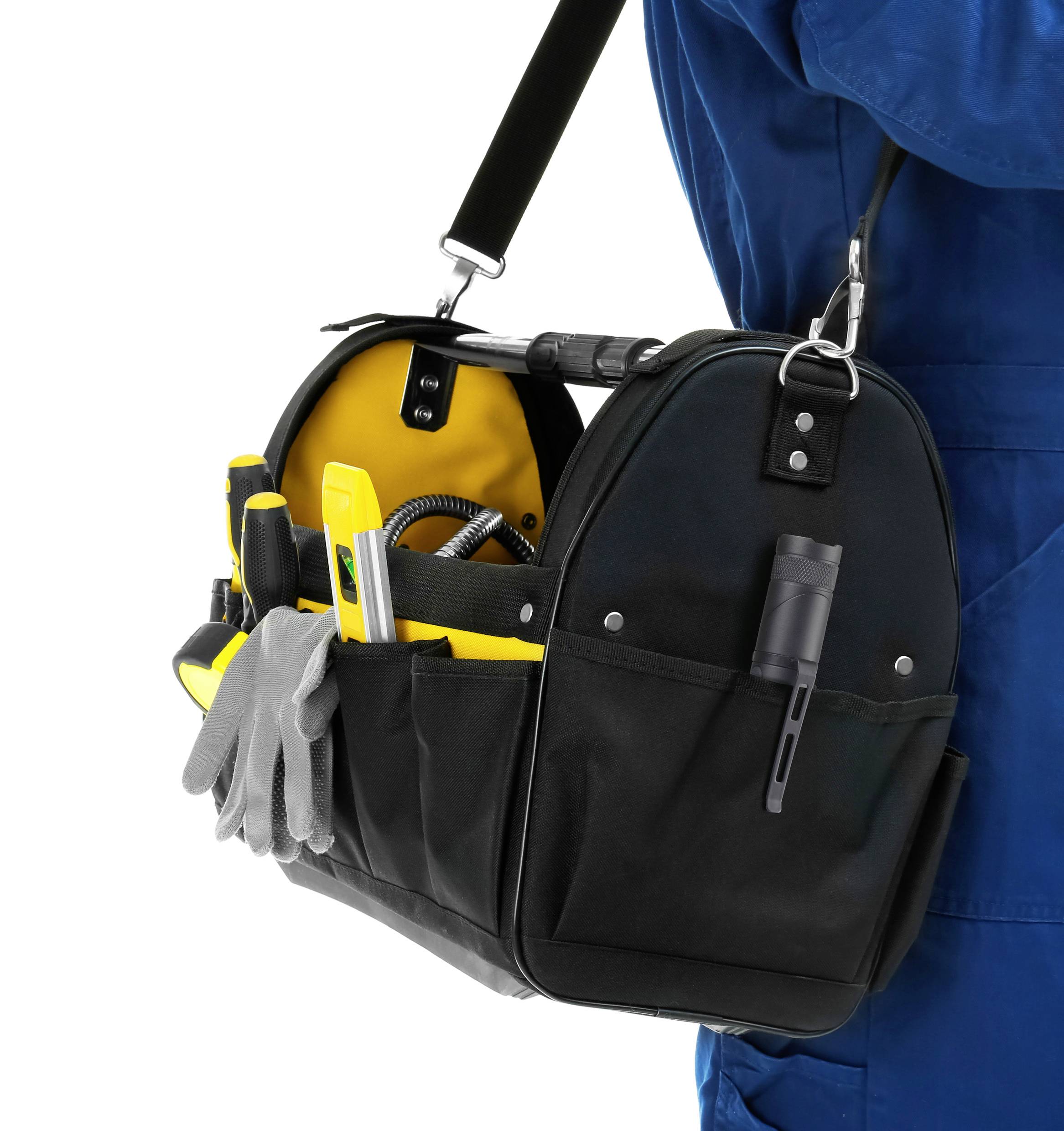 Eine schwarze Werkzeugtasche aus Stoff, gefüllt mit verschiedenen Werkzeugen wie Schraubendreher und Zangen, hängt über der Schulter einer Person in blauer Arbeitskleidung.