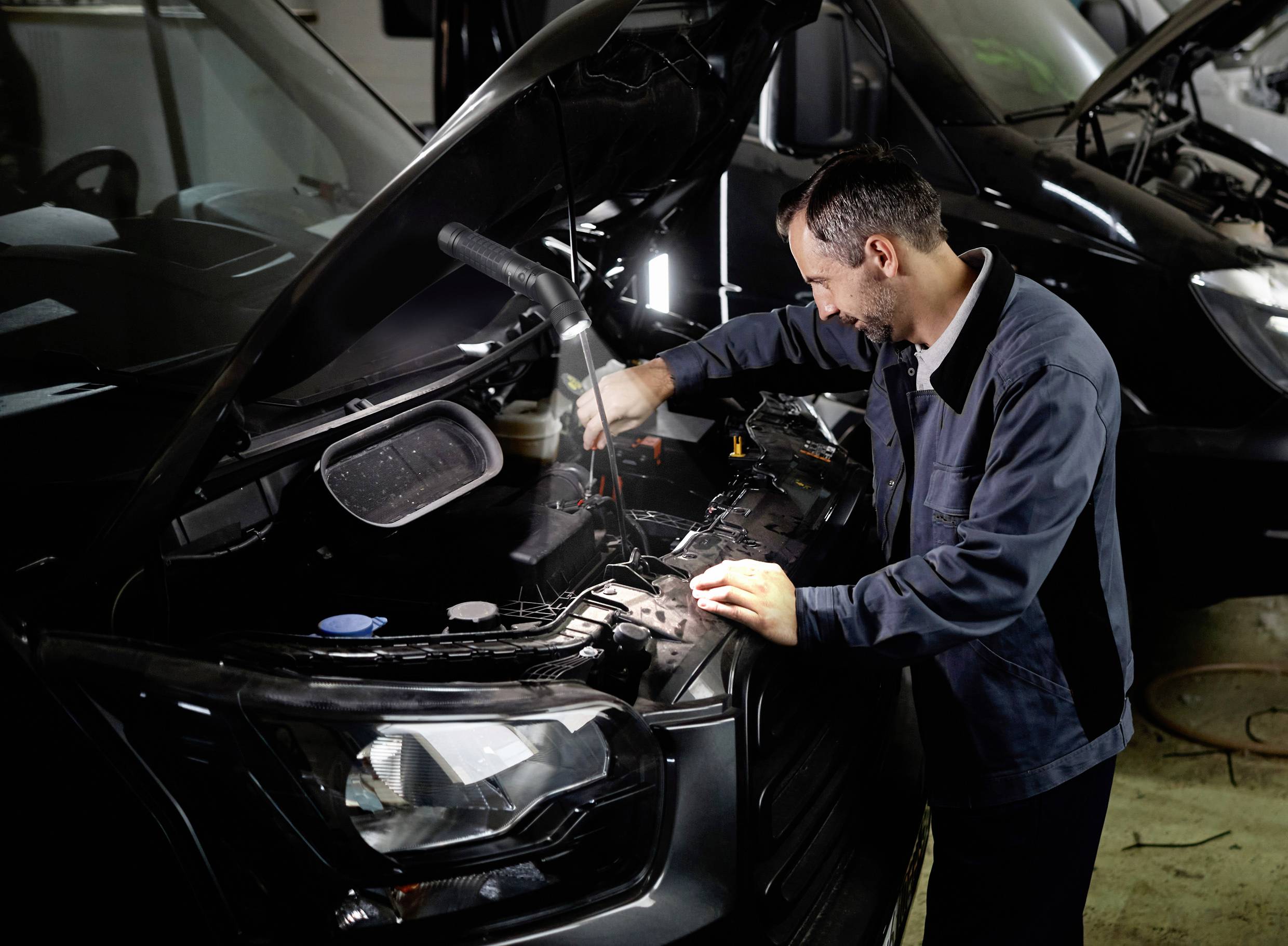 Ein Automechaniker arbeitet unter der geöffneten Motorhaube eines Fahrzeugs in einer Werkstatt, während er mit Werkzeugen hantiert.