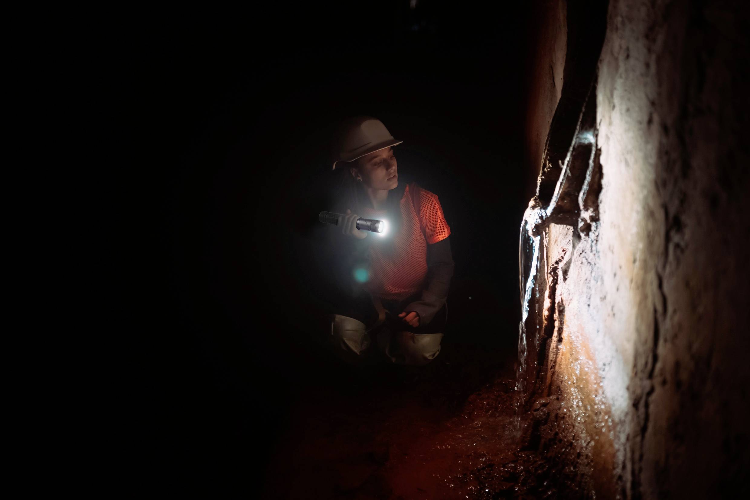 Eine Person mit Helm und Taschenlampe untersucht eine dunkle Höhle oder einen Tunnel, beleuchtet die feuchten Wände aufmerksam.