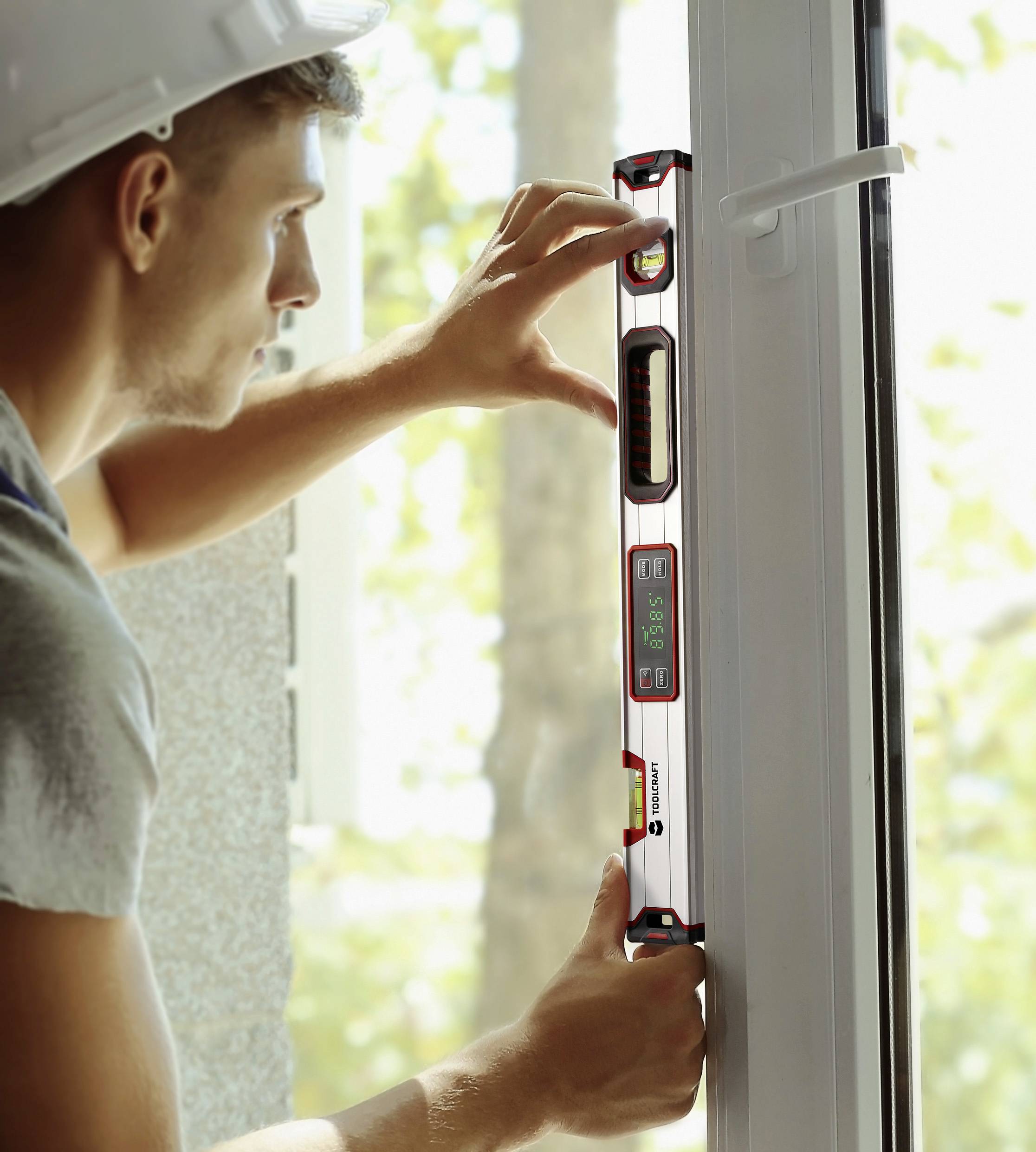 Ein Mann hält eine Wasserwaage an einem Fensterrahmen, um die Ausrichtung zu überprüfen. Im Hintergrund sind Bäume sichtbar.