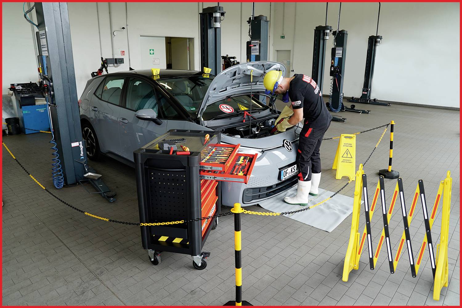 Ein Mechaniker steht neben einem Auto in einer Werkstatt und arbeitet am Motorraum. Sicherheitsabsperrungen und Werkzeuge sind sichtbar.