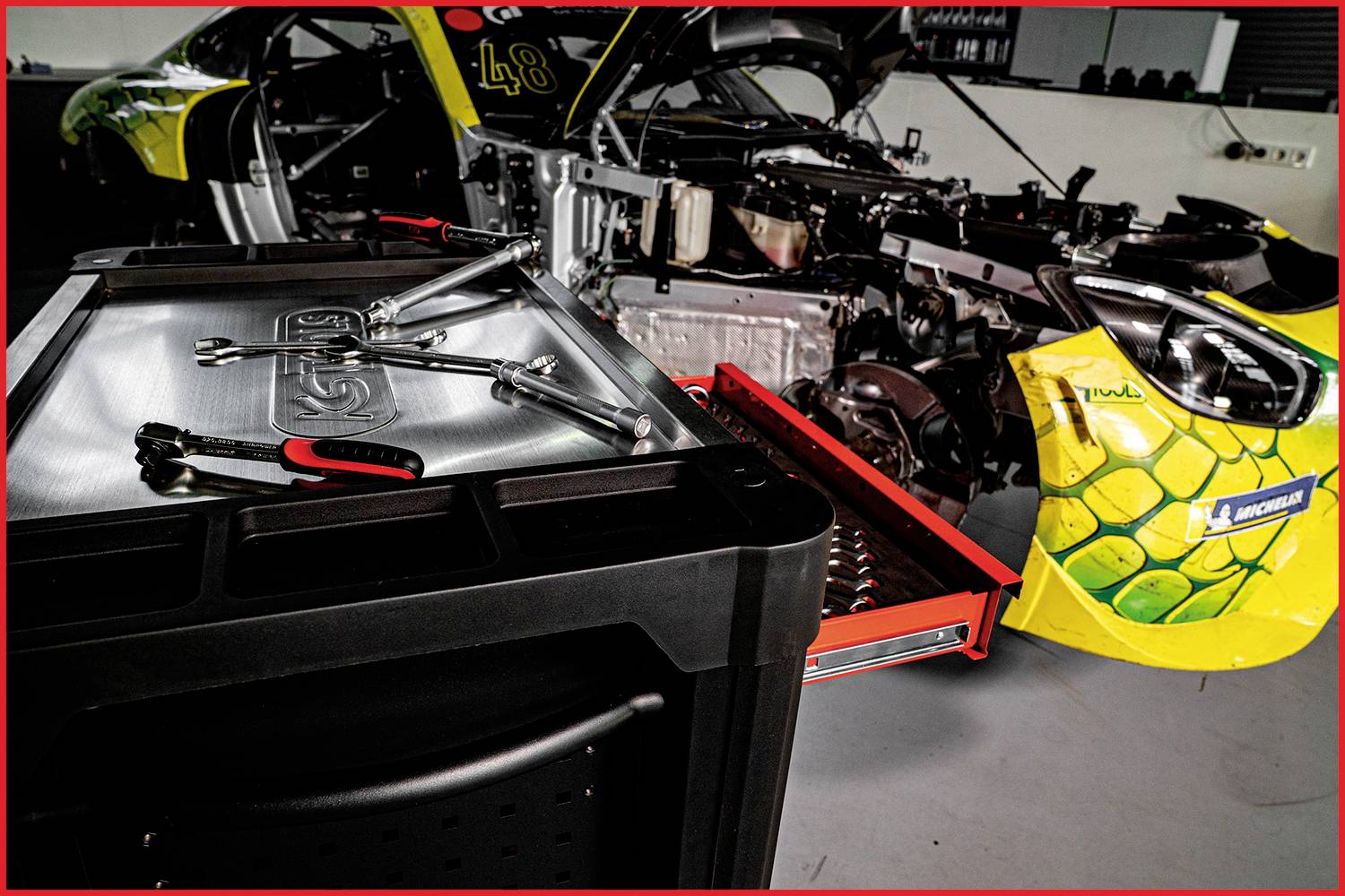 Ein Rennwagen steht in einer Werkstatt mit geöffneter Motorhaube. Auf einem Wagen liegen Werkzeuge. Im Hintergrund sind Autoteile sichtbar.