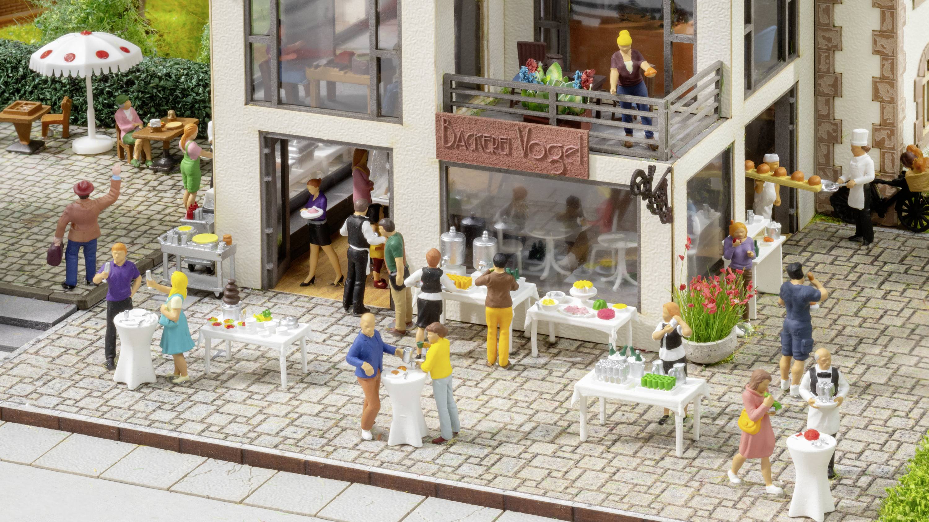 Ein Modell eines Straßencafés mit Miniaturfiguren, die Getränke und Speisen servieren. Kunden stehen an weißen Tischen im Freien.