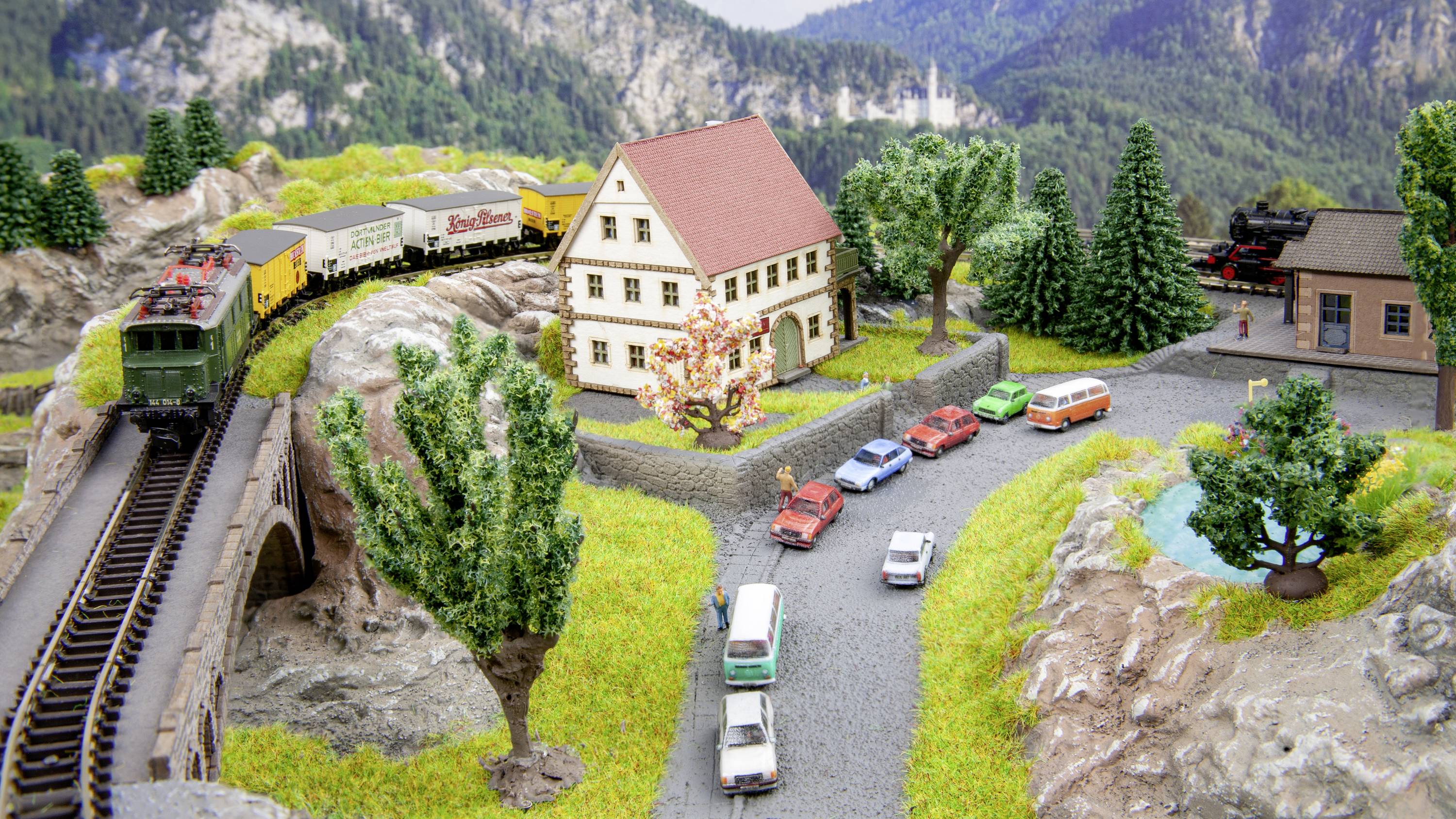 Modelleisenbahn mit Zug auf Brücke, Landschaft mit Hügeln, Bäumen, Häusern und parkenden Autos. Hintergrund: Berge und bewaldete Umgebung.