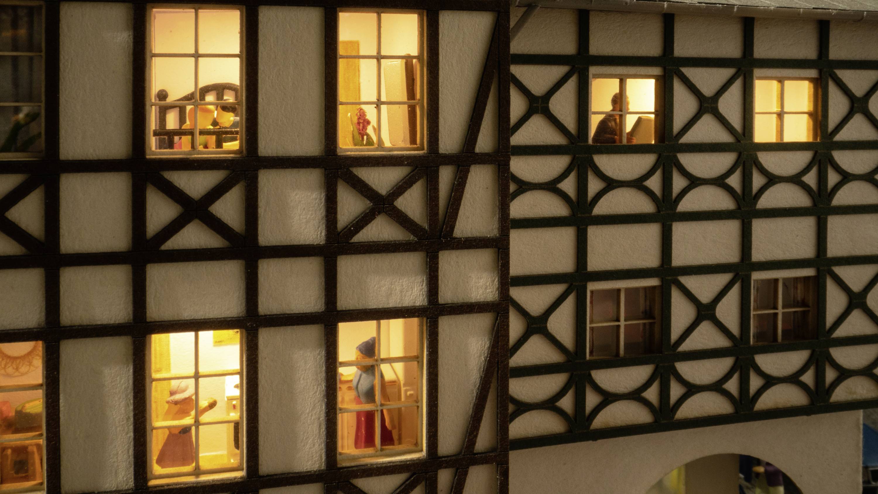 Beleuchtete Fachwerkhäuser bei Nacht, Fenster zeigen Innenräume mit Figuren, die Alltagsszenen darstellen.