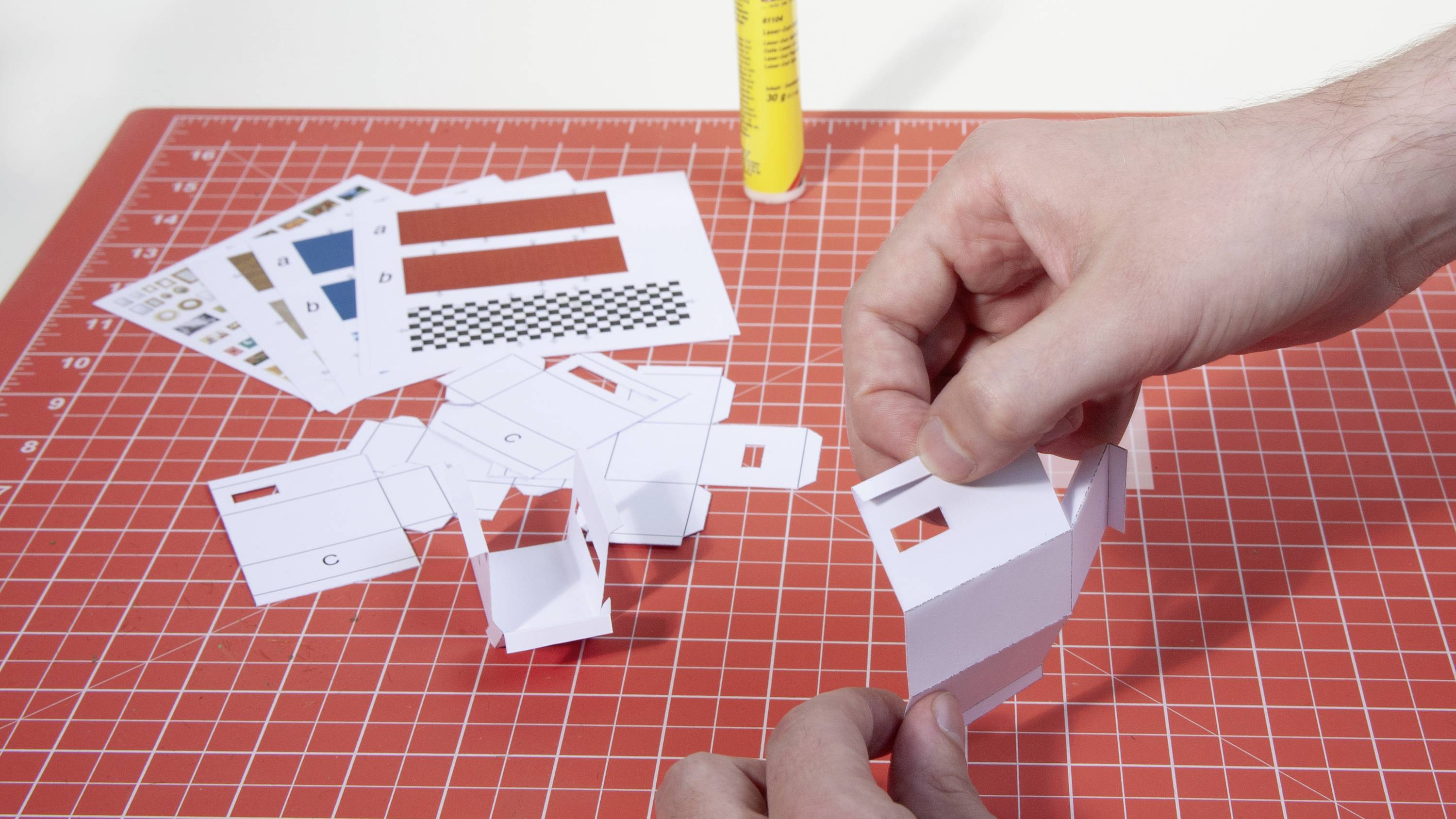 Zwei Hände falten Papiermodelle auf einer Schneidematte mit ausgeschnittenen farbigen Formen und einer gelben Kleberflasche.