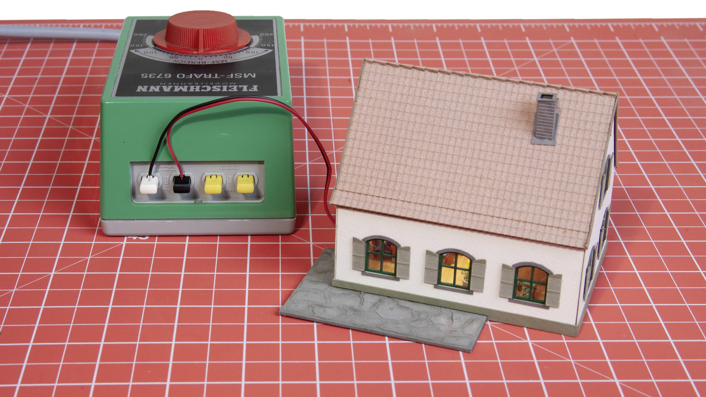 Ein kleines Modellhaus steht auf einer rot karierten Oberfläche neben einem grünen Elektroschalter mit Kabelverbindungen.
