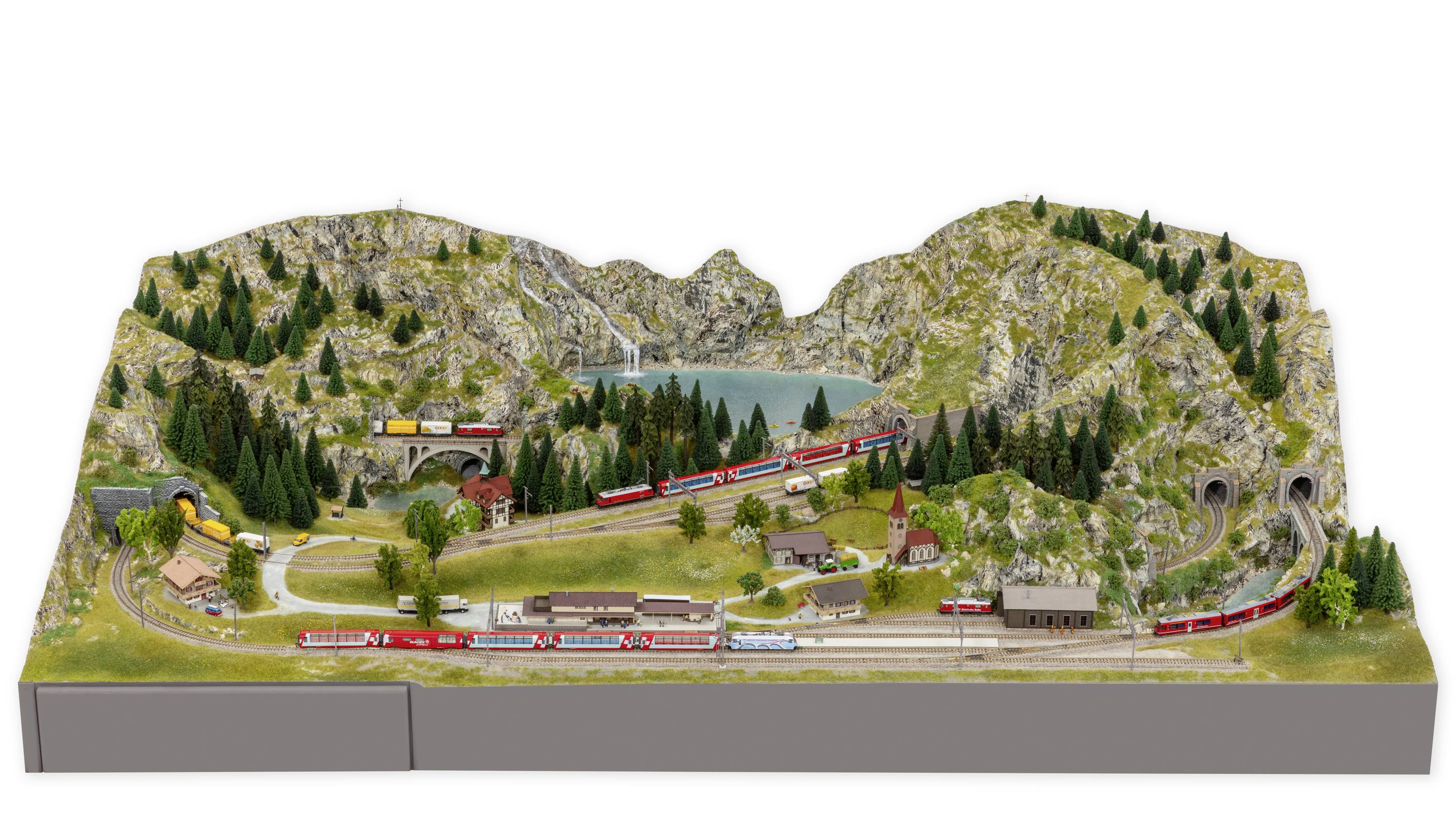 Modelleisenbahn-Diorama zeigt eine alpine Landschaft mit Bergen, Seen, Tunneln und Zügen. Die Szenerie umfasst kleine Häuser und Wälder.
