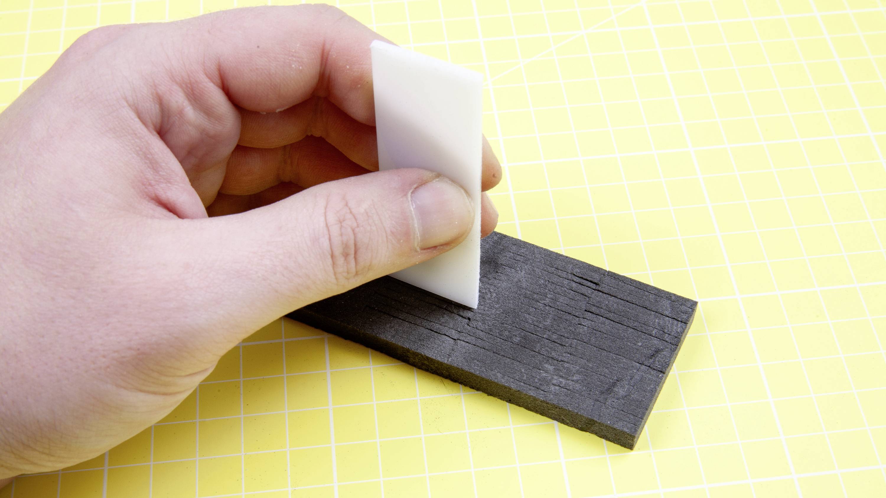 Eine Hand hält eine weiße Klinge und schneidet ein schwarzes rechteckiges Material auf einem gelben Rasterhintergrund.