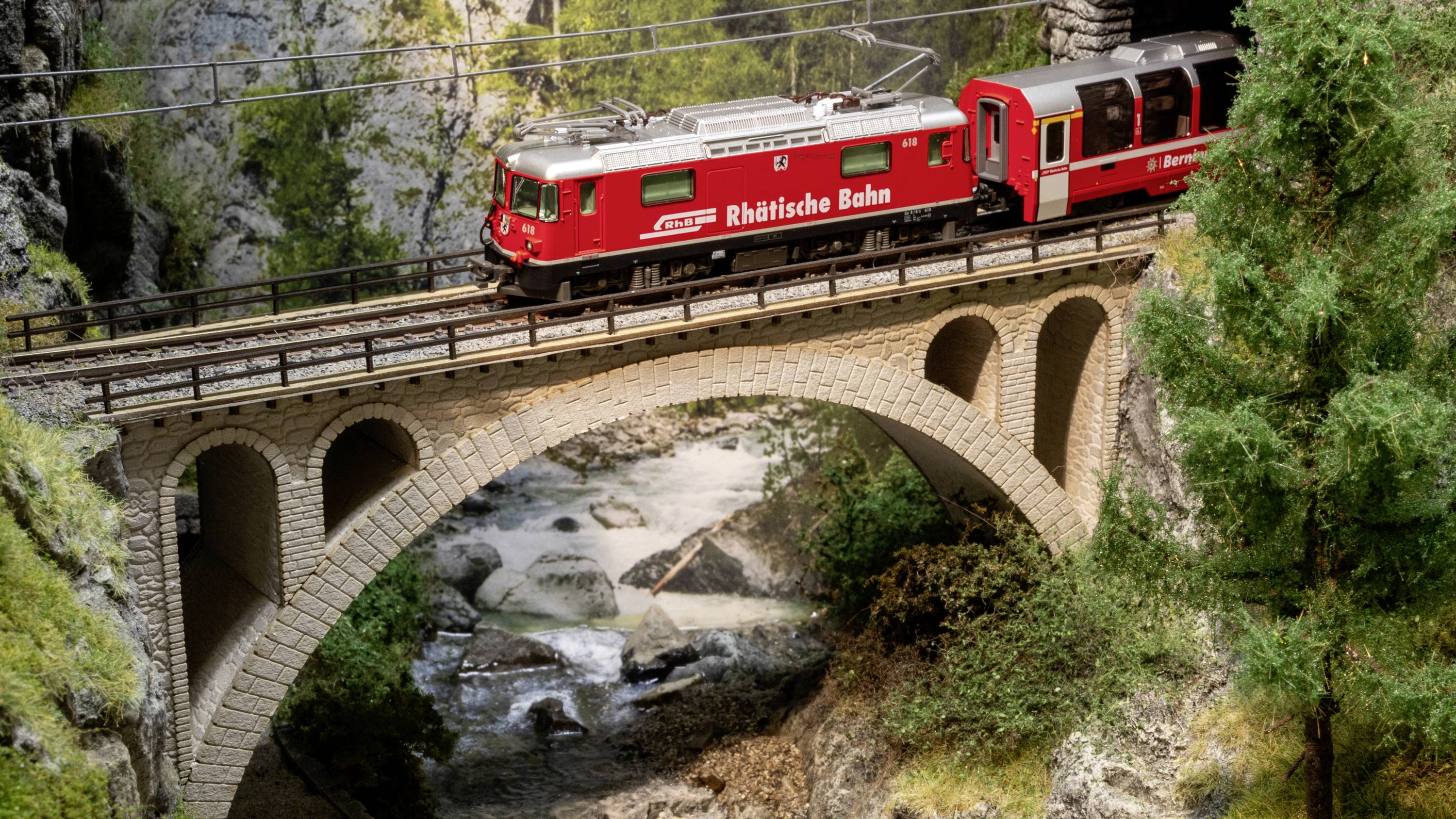 Eine rote Rhätische Bahn-Lokomotive fährt über eine steinerne Brücke in einer bergigen Landschaft mit dichter Vegetation.