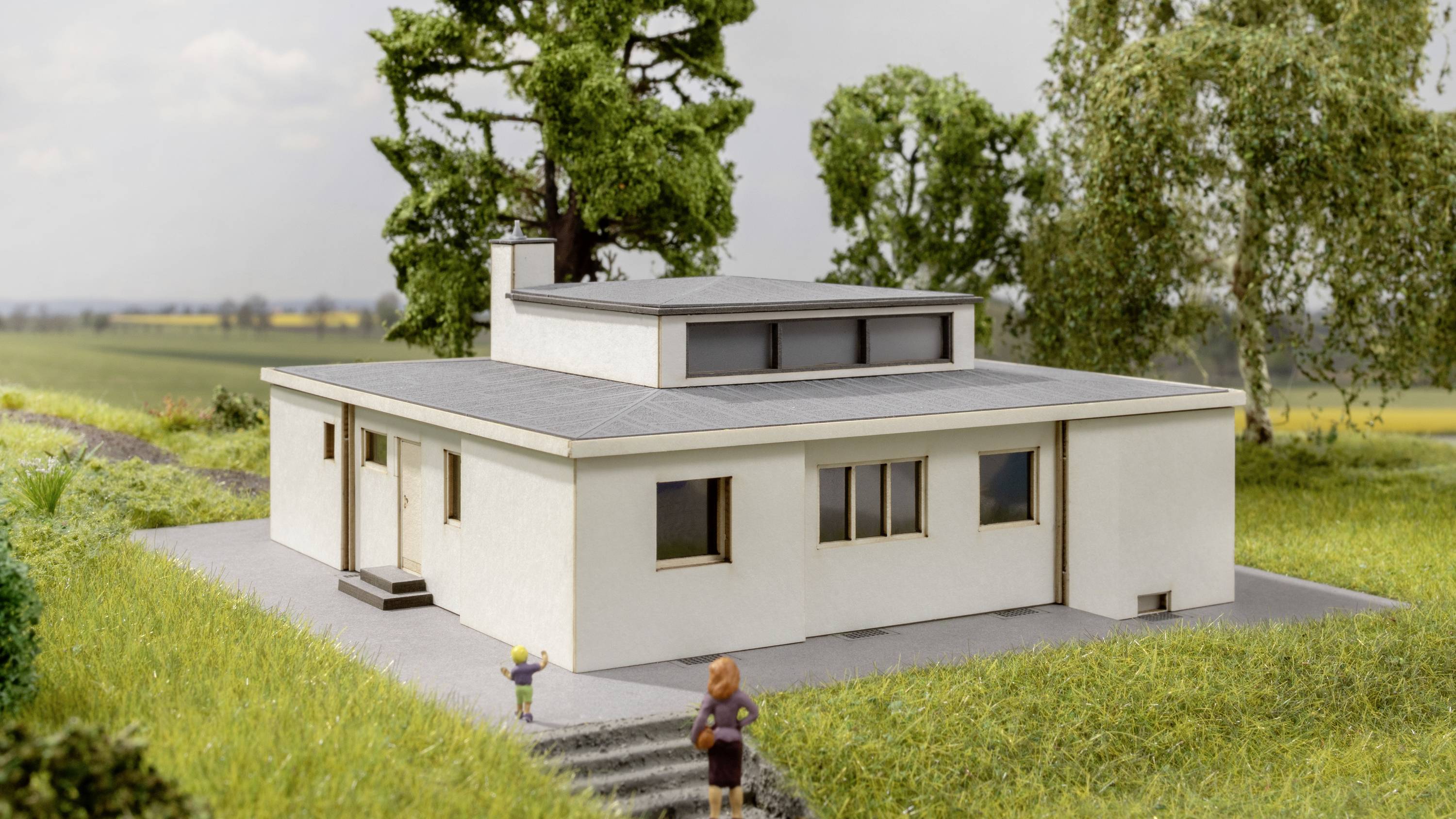 Ein modernes weißes Flachdachhaus steht auf einer grasbewachsenen Fläche. Im Vordergrund ist eine Frau, die auf das Haus zugeht.