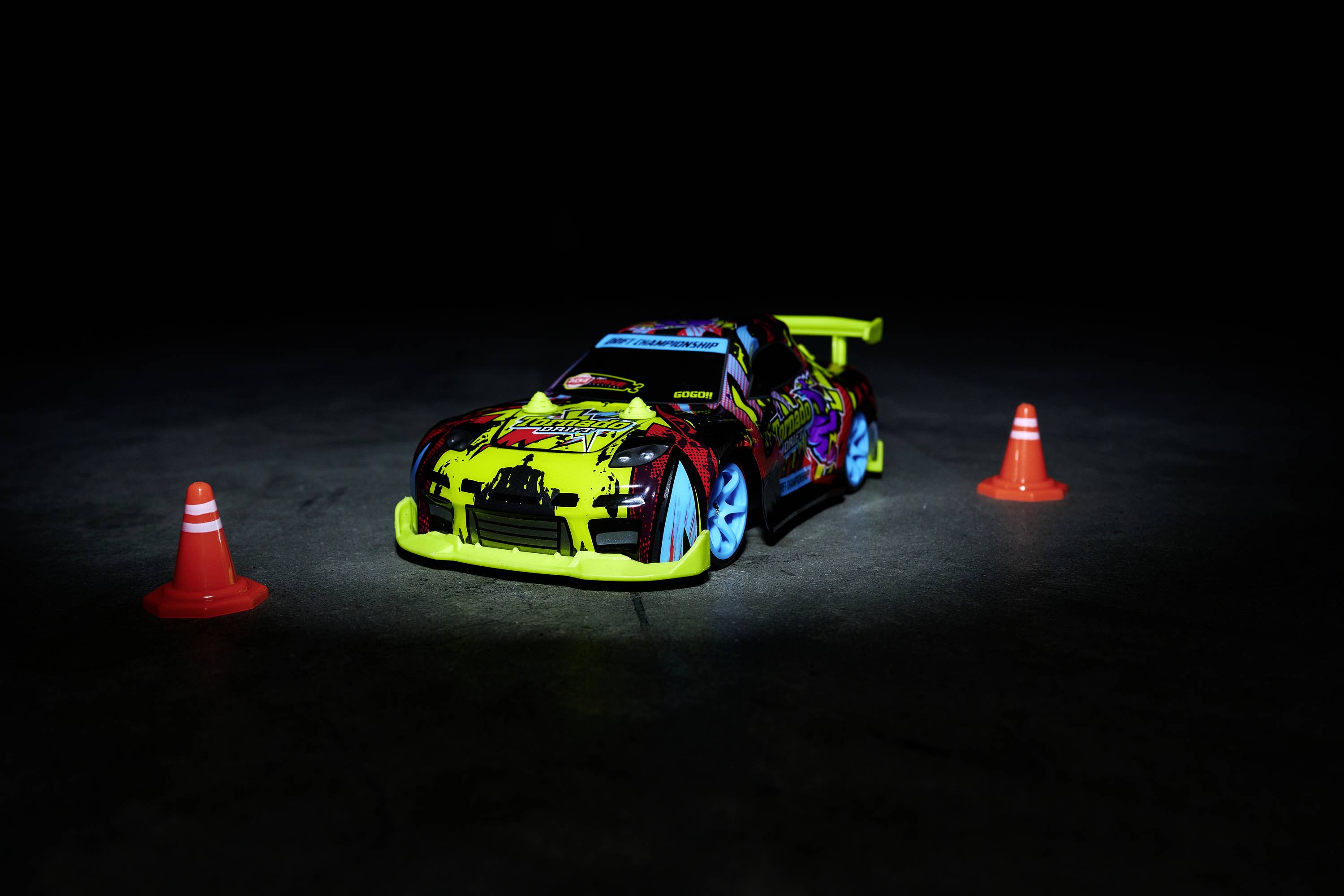 Spielzeugauto mit buntem Design und großen Spoilern auf dunklem Boden zwischen zwei kleinen orangenen Verkehrskegeln.
