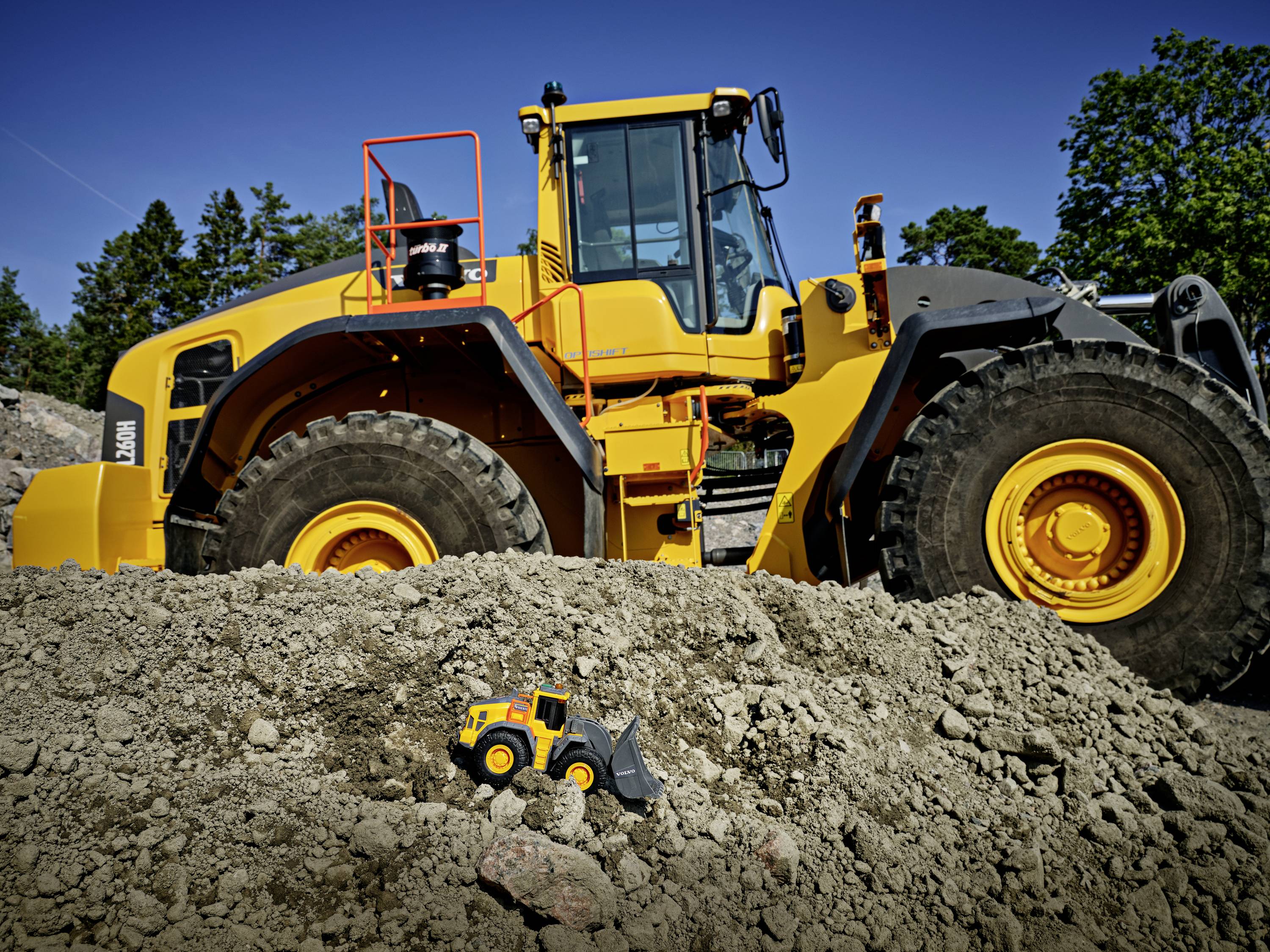 Ein großer gelber Bagger steht auf einer Baustelle, während im Vordergrund ein Spielzeugbagger im Sand zu sehen ist.