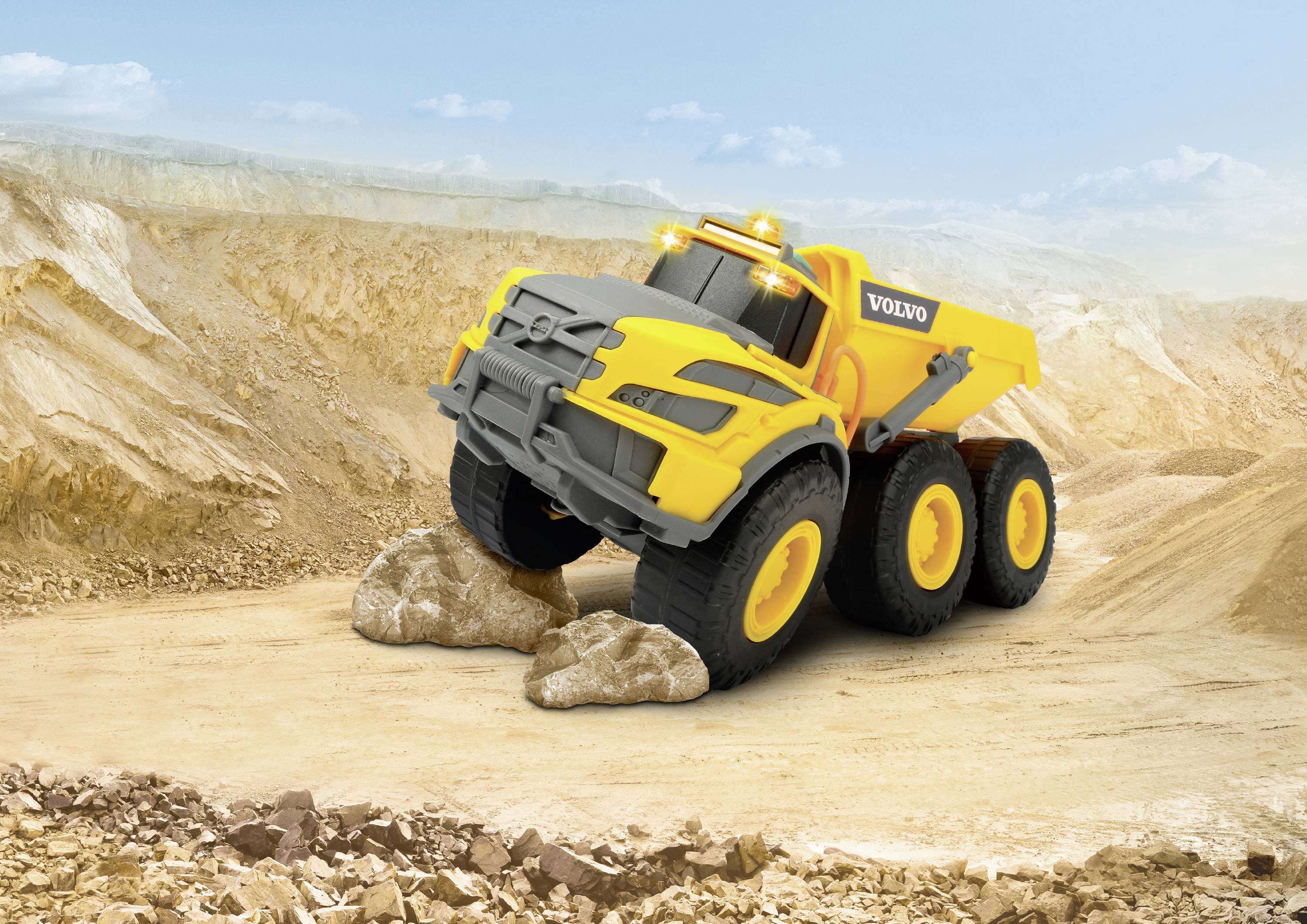 Ein gelber Spielzeug-Muldenkipper fährt auf einer Baustelle über zwei Felsen. Sandige Hügel sind im Hintergrund zu sehen.