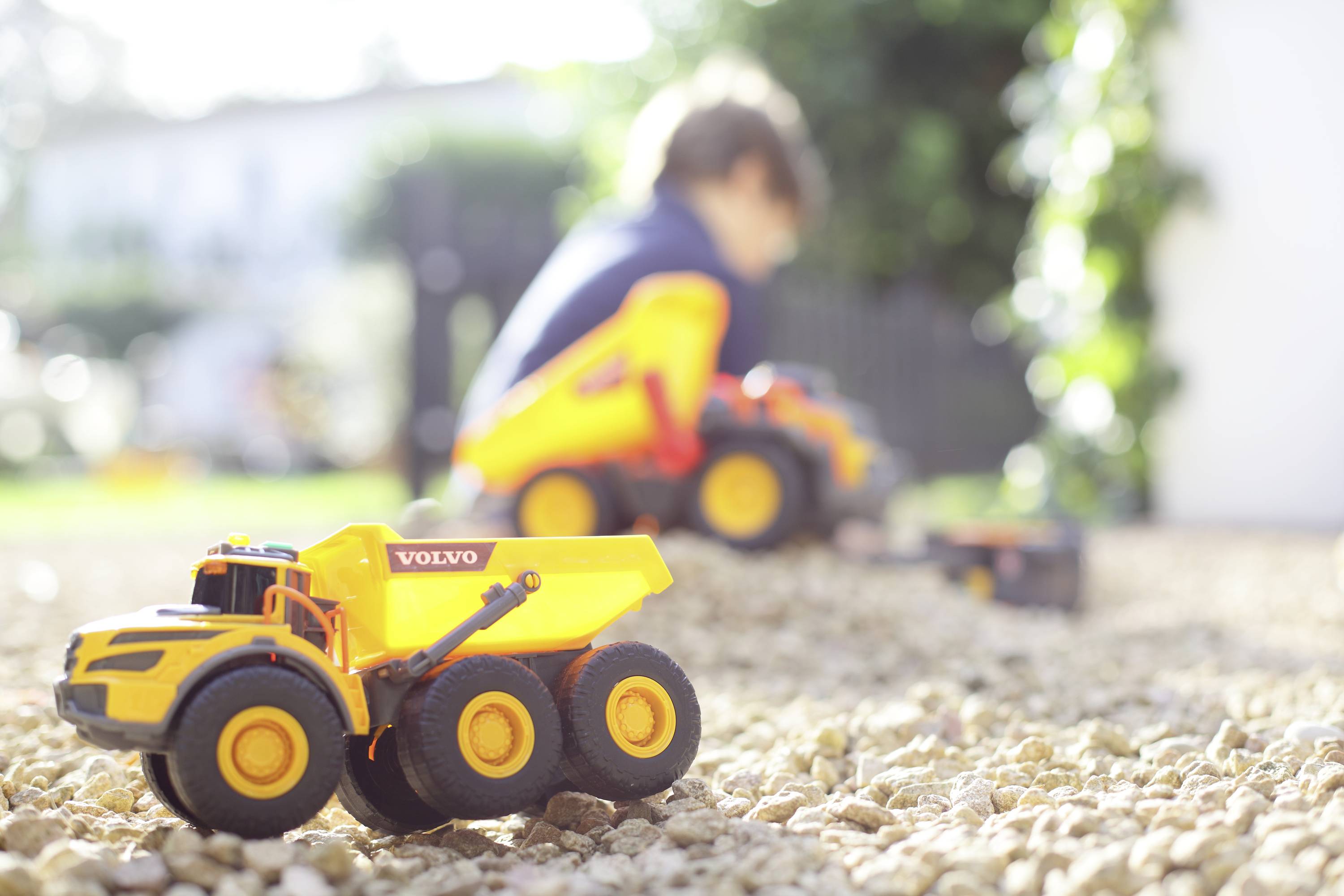 Ein gelber Spielzeugkipper steht scharf im Vordergrund auf Kies. Im Hintergrund spielt ein Kind unscharf mit einem ähnlichen Fahrzeug im Garten.