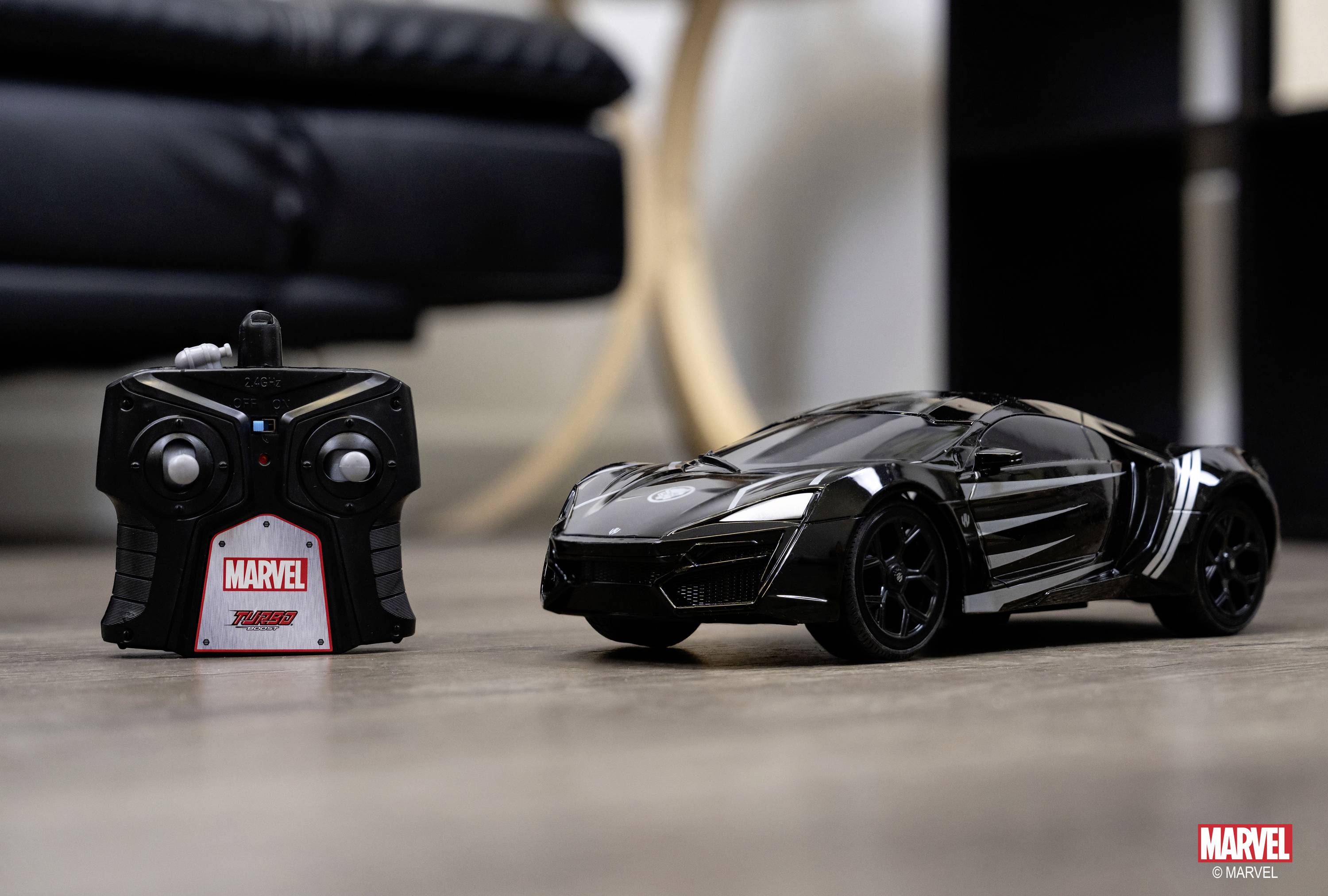 Ein ferngesteuertes Spielzeugauto in Schwarz mit einem Controller, auf einem Holzboden stehend. Im Hintergrund unscharfe Möbeldetails.