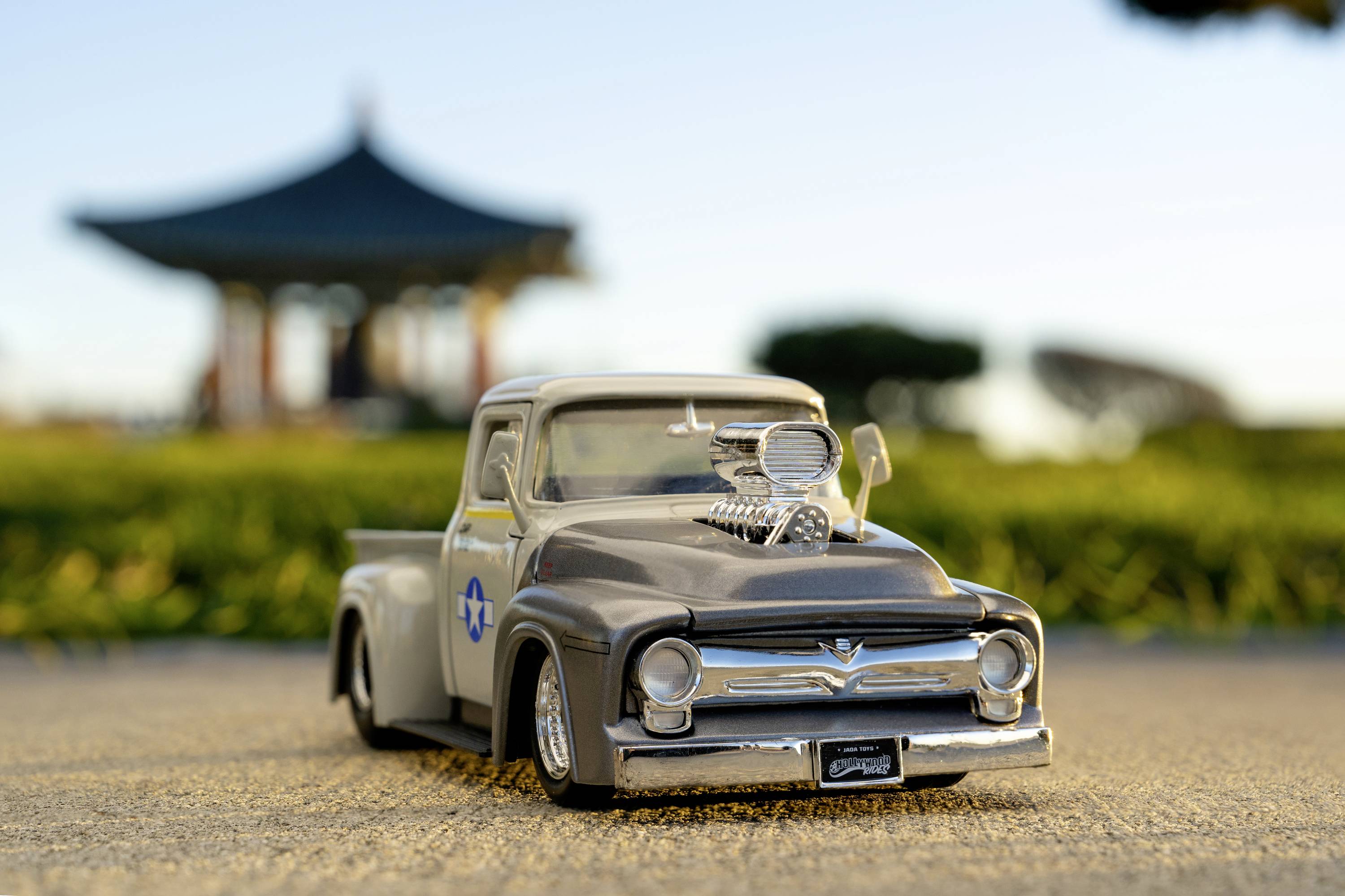 Ein graues, maßstabsgetreues Modell eines Oldtimer-Pickups steht im Fokus, im Hintergrund ein Pavillon und grüne Landschaft.