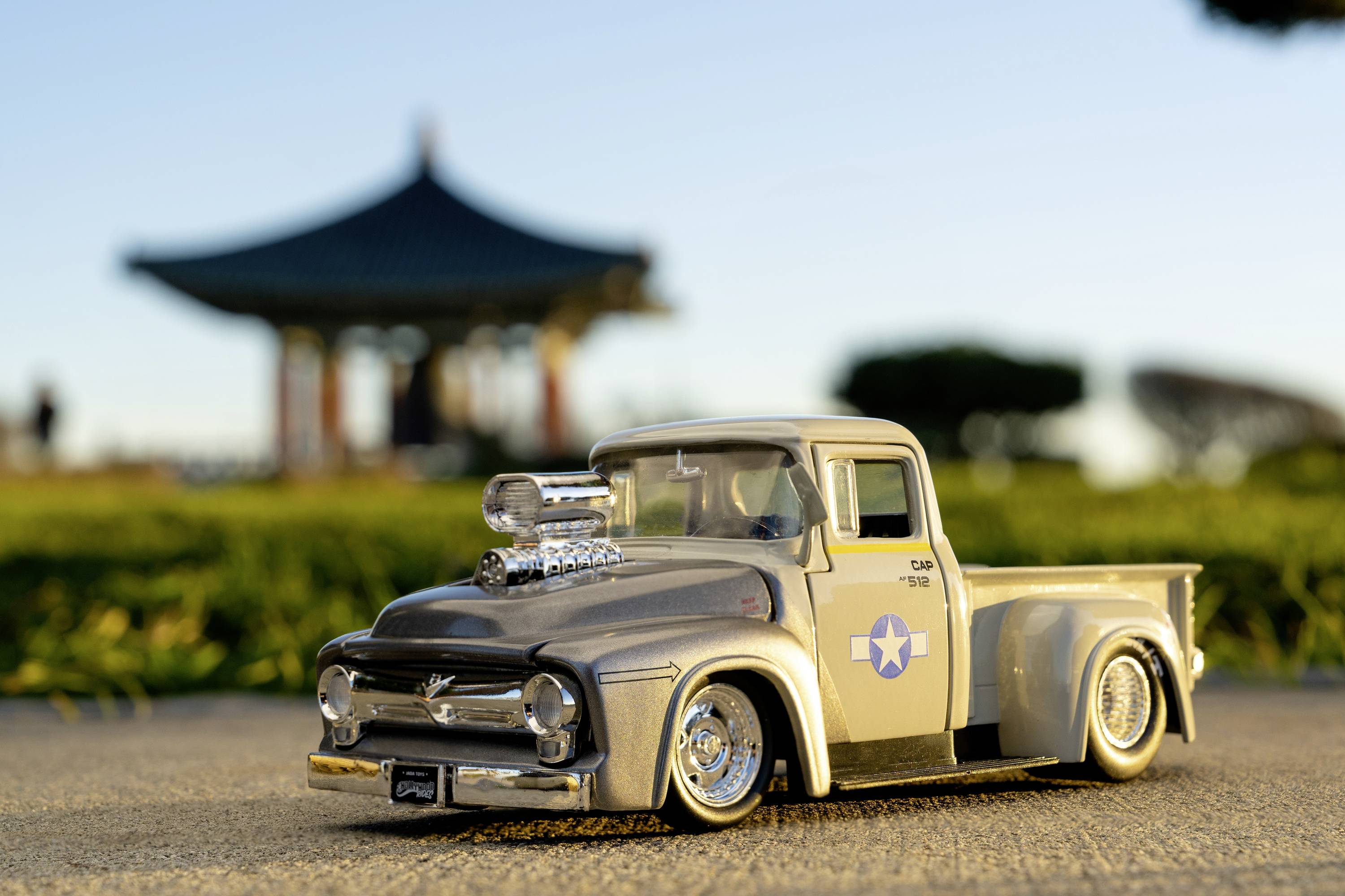 Kleines Spielzeugmodell eines Vintage-Pickups fährt auf einer sonnigen Straße mit einem unscharfen Pavillon im Hintergrund.