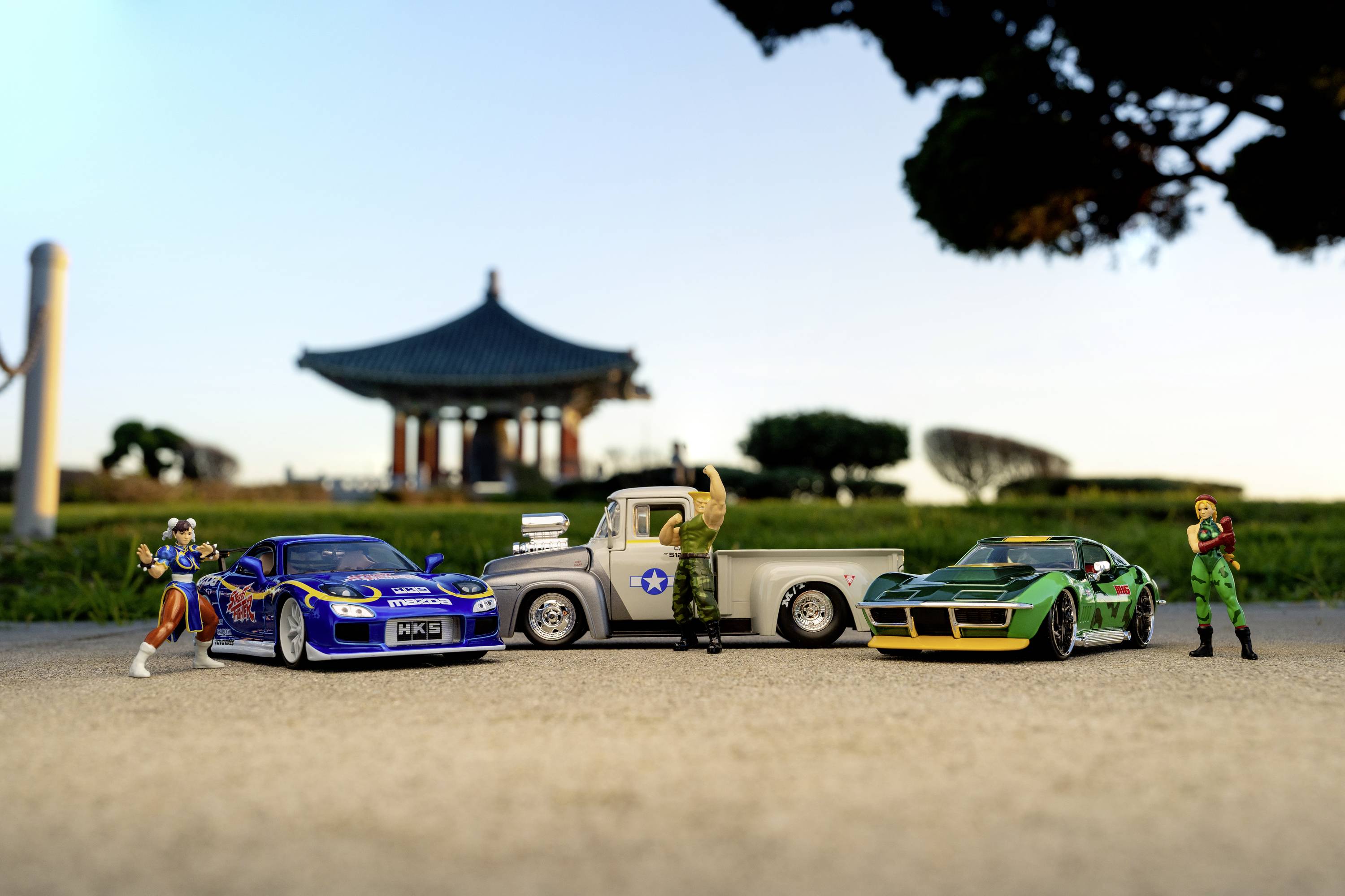 Spielzeugautos und Actionfiguren stehen nebeneinander auf einem Bürgersteig. Im Hintergrund ist ein Pavillon vor einem blauen Himmel sichtbar.