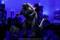 Eine schwarze Godzilla-Figur mit rosa Stacheln steht vor einer Kulisse aus verschwommenen Hochhäusern. Links unten liegt ein kleiner Controller.