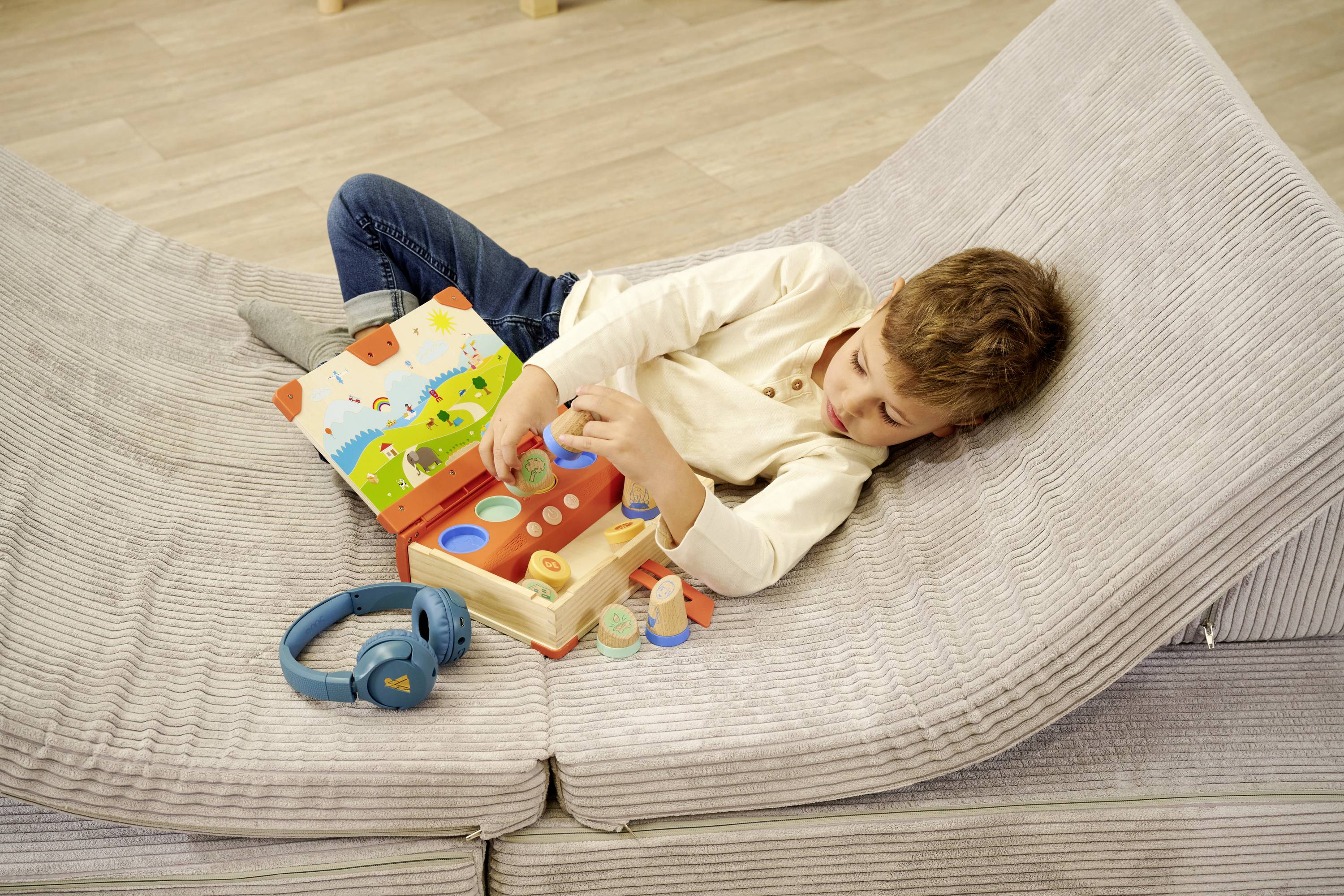 Ein Kind liegt auf einem grauen Sofa und spielt mit einem Holzspielzeug. Ein offenes buntes Buch und blaue Kopfhörer liegen daneben.