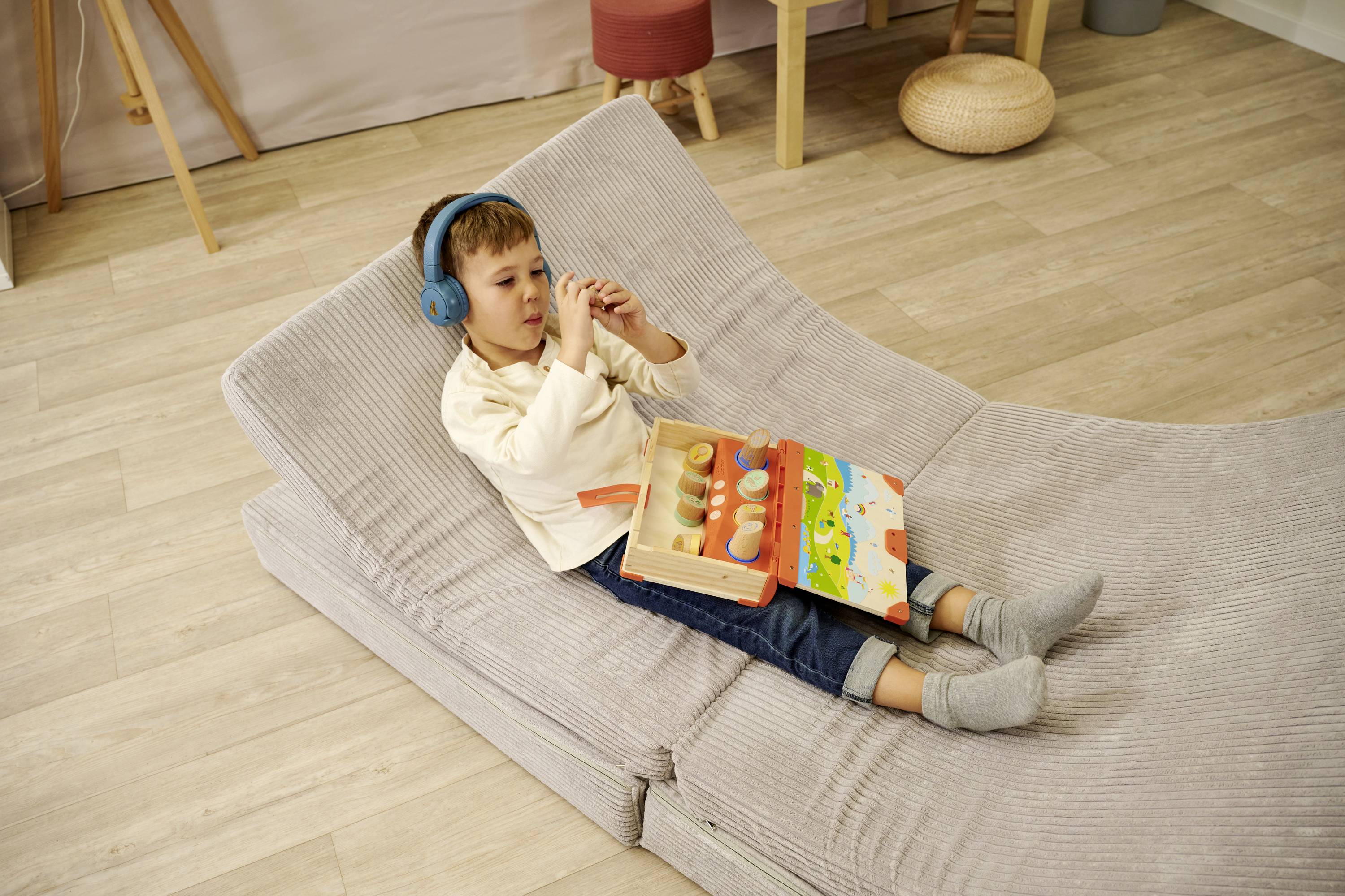 Ein Kind liegt auf einem grauen Polster, trägt blaue Kopfhörer und spielt mit einem Holzspielzeug in einem Kinderzimmer.