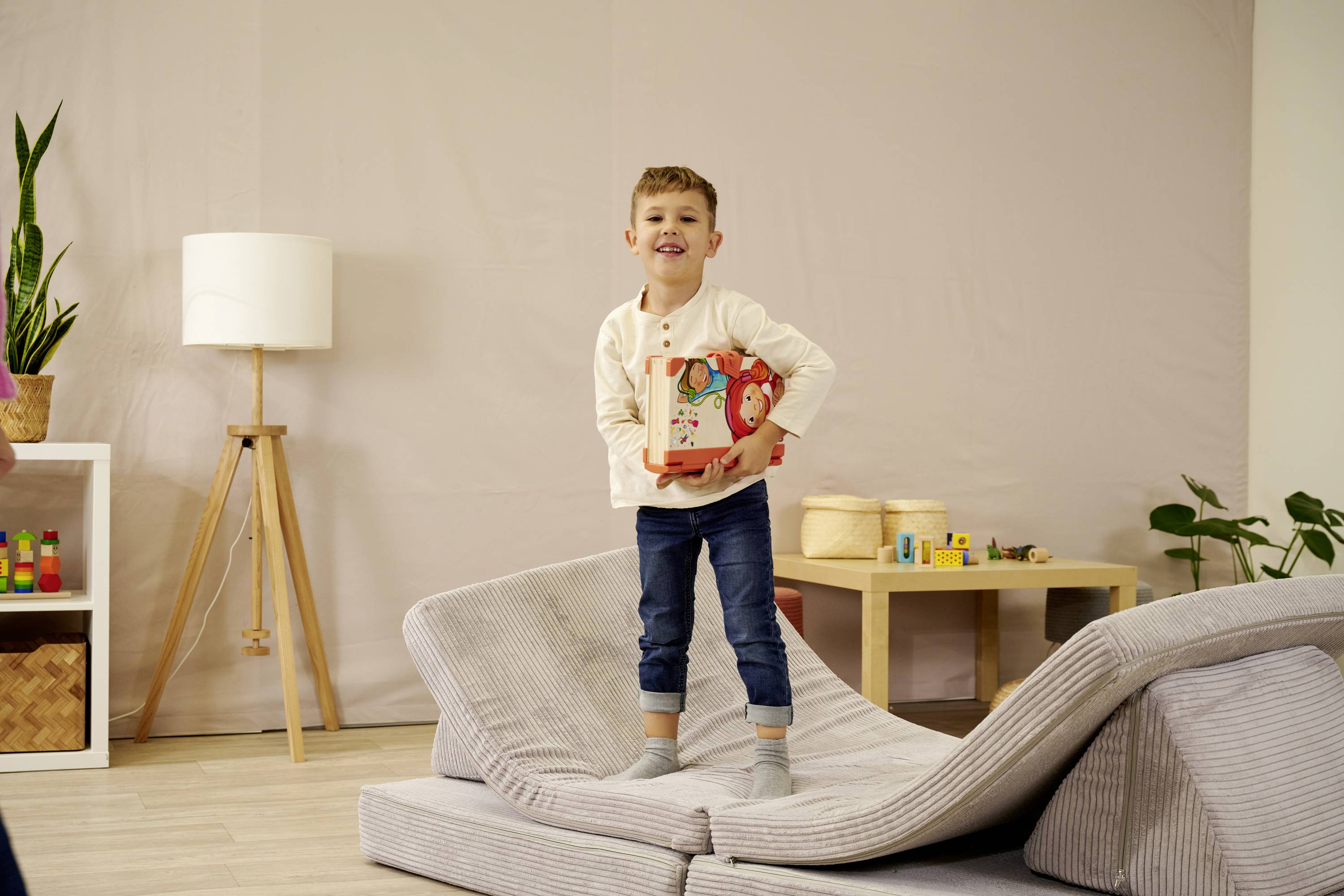 Ein lächelnder Junge steht auf einem grauen Sofa und hält einen Spielzeugbus. Im Hintergrund sind Pflanzen und eine Stehlampe zu sehen.
