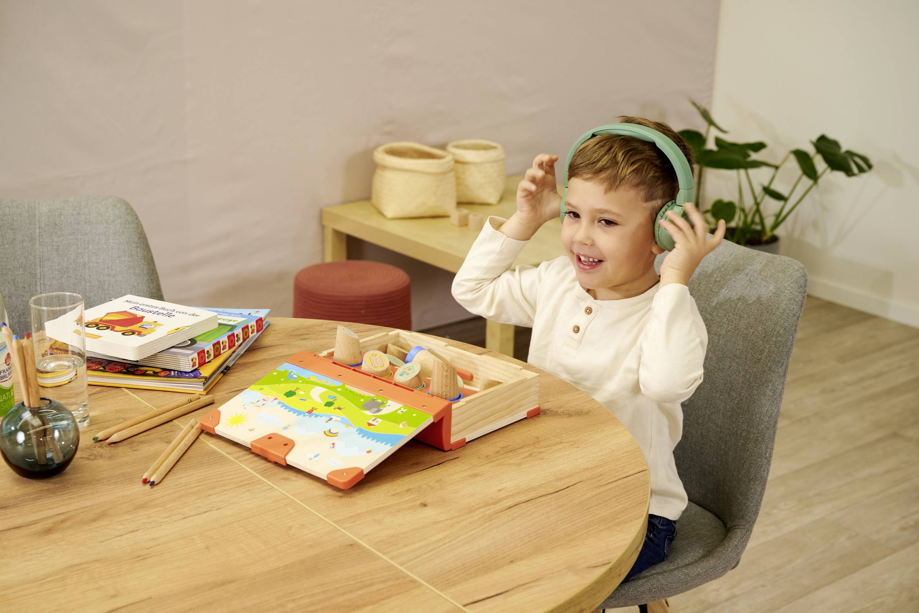 Ein Kind sitzt an einem Tisch und trägt Kopfhörer. Vor ihm liegen Kinderbücher und Holzspielzeug. Im Hintergrund sind Pflanzen sichtbar.