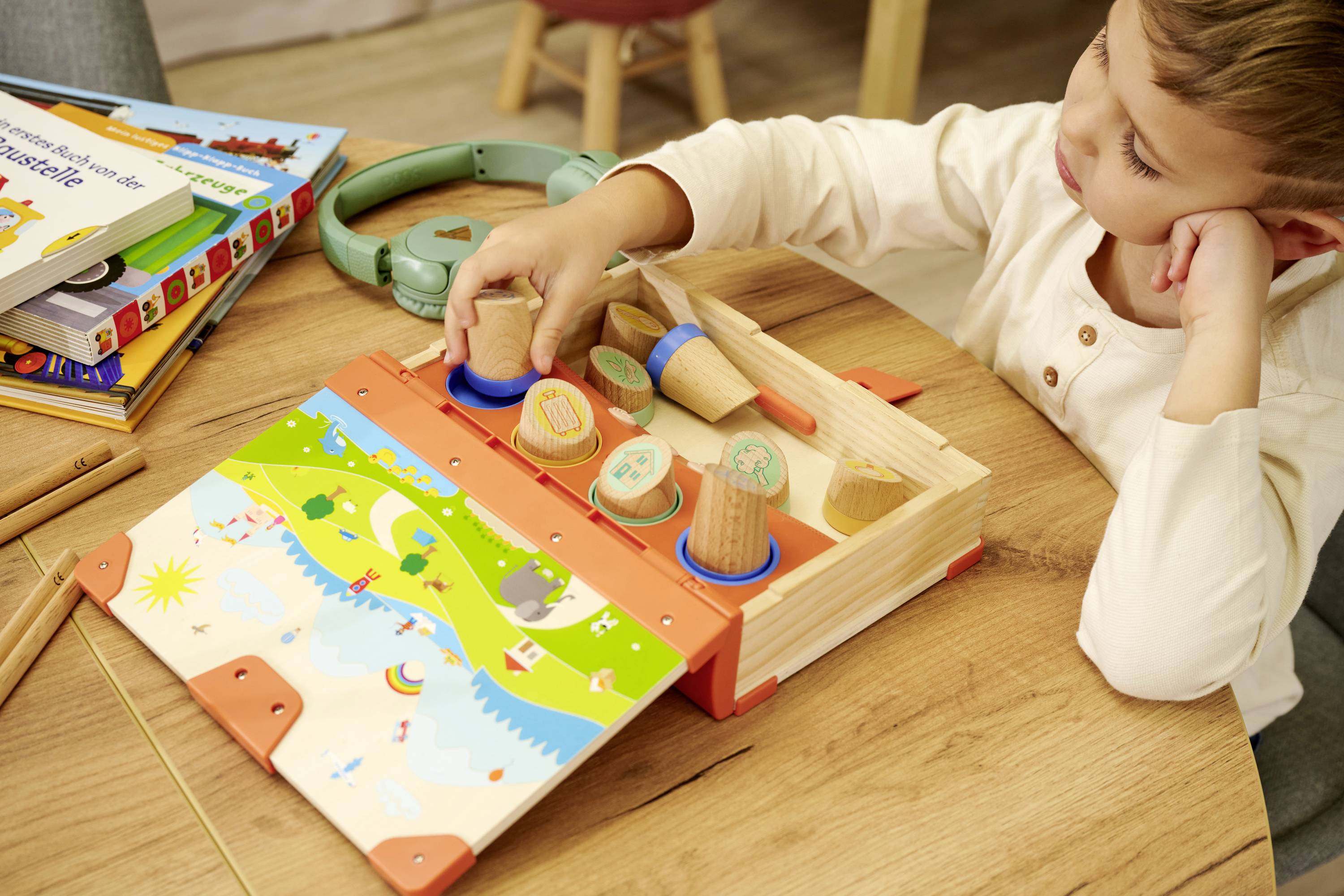 Kind spielt mit einem Holzpuzzle am Tisch. Zahlreiche Holzstücke passen in eine bunte Karte. Bücher und Kopfhörer im Hintergrund.