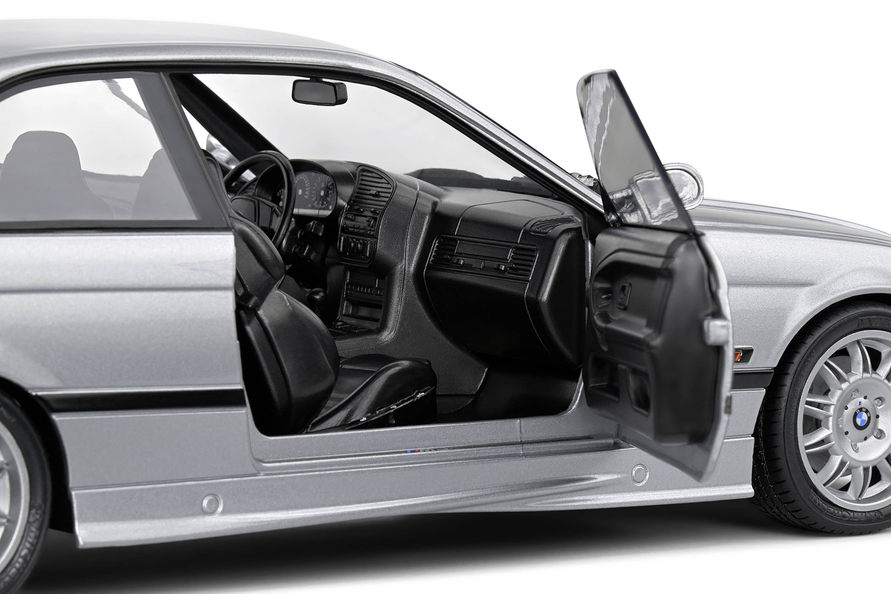 Silbernes Auto mit geöffneter Fahrertür. Der Blick zeigt ein detailliertes, ledernes Interieur mit Sicht auf das Lenkrad und das Armaturenbrett.