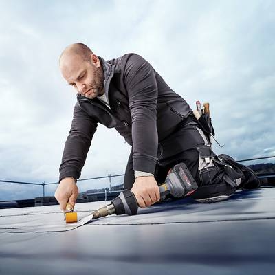 Ein Mann auf einem Dach arbeitet mit Werkzeugen an einer Abdeckung. Er trägt Arbeitskleidung und ist in einer knienden Position.
